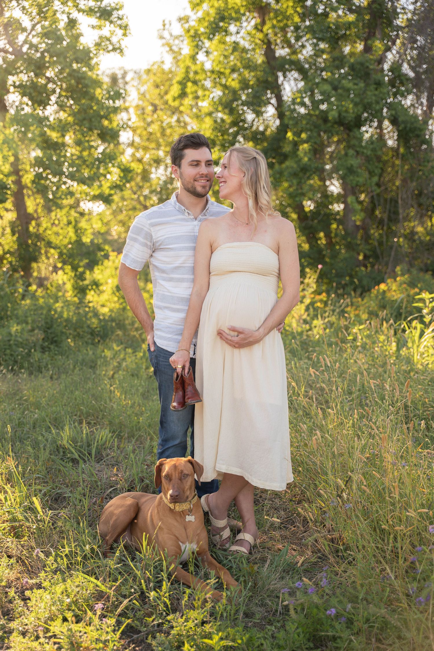 Pregnant woman in yellow dress with partner and brown dog in sunny outdoor maternity photo session.