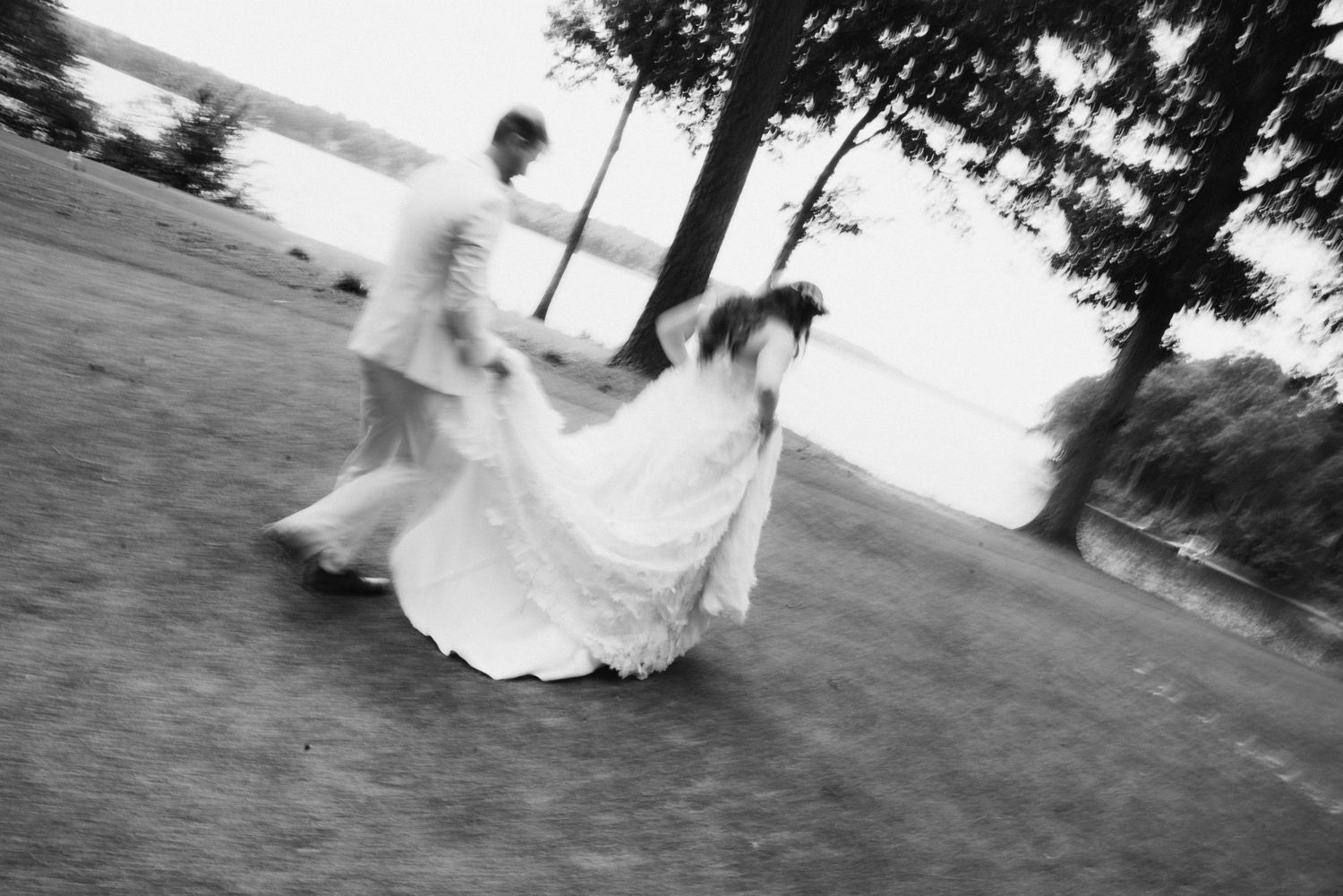 Black and white motion blur photo of bride and groom dancing outdoors near a lakeside venue.
