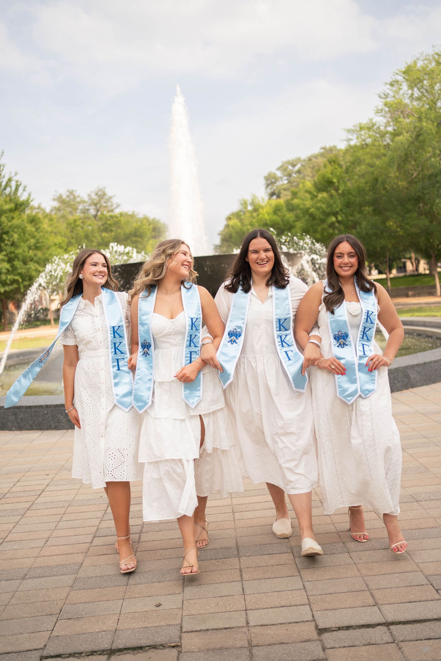 Four Kappa Kappa Gamma sorority graduates in white dresses with blue KKT stoles walking by fountain.