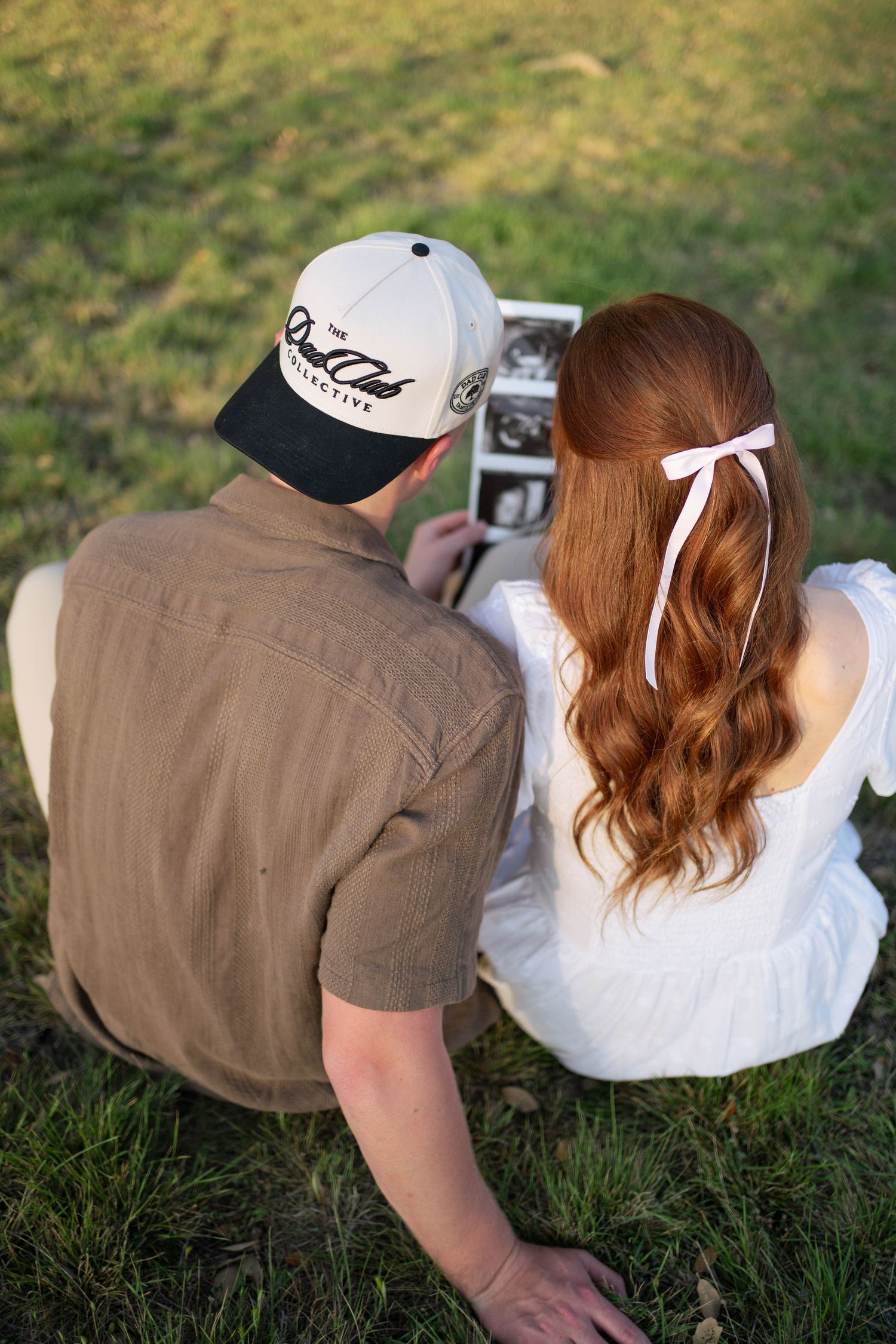 Couple sitting on grass viewing ultrasound photos, woman with red hair and white bow, man in brown shirt and cap.