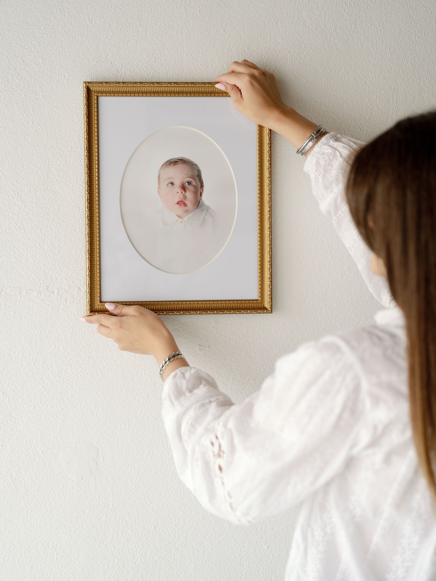 A woman in a white blouse hangs a gold-framed oval portrait of a baby on a white wall.