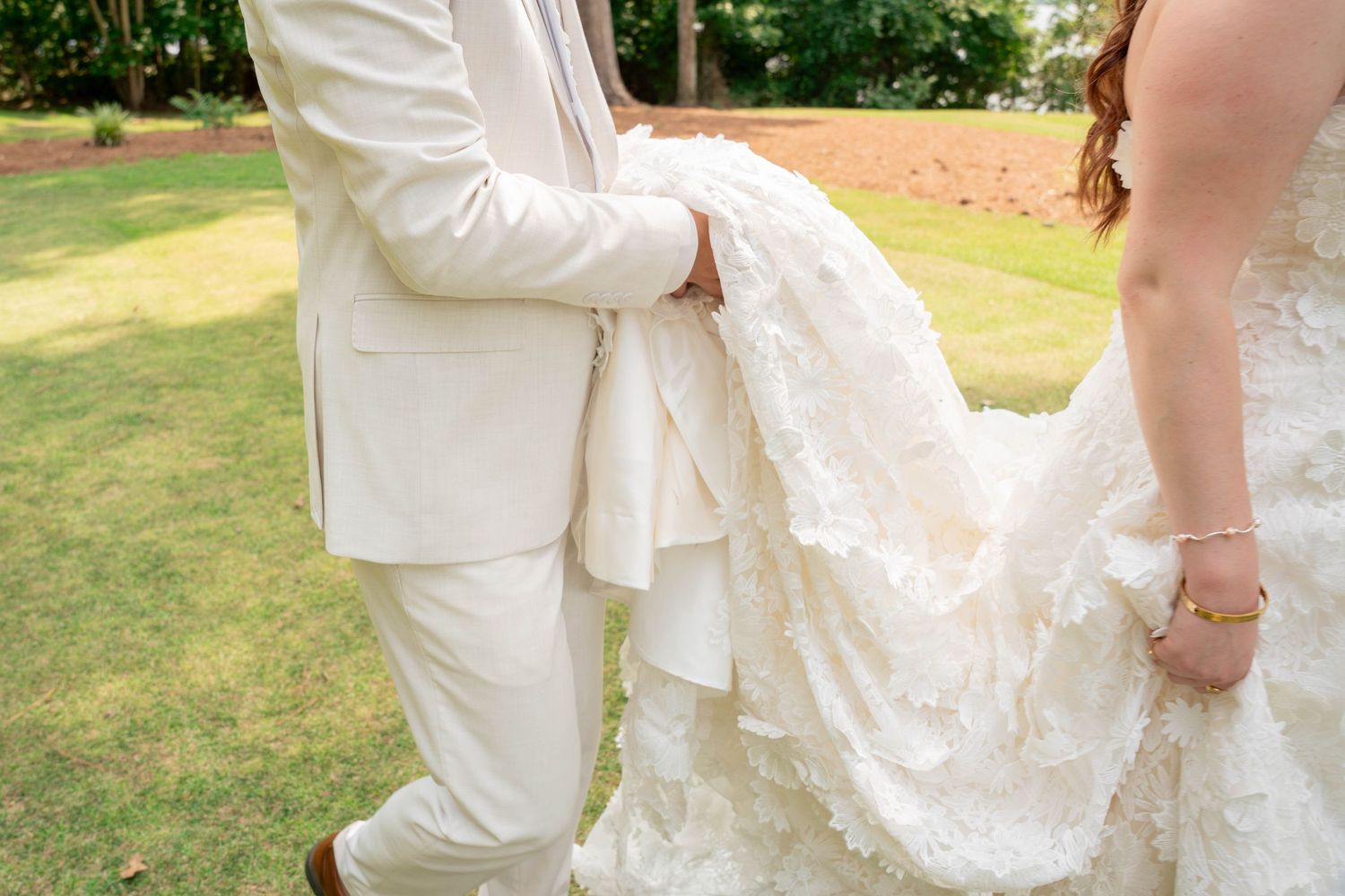 Groom in white suit carries bride's lace wedding dress train on green lawn at outdoor wedding ceremony.