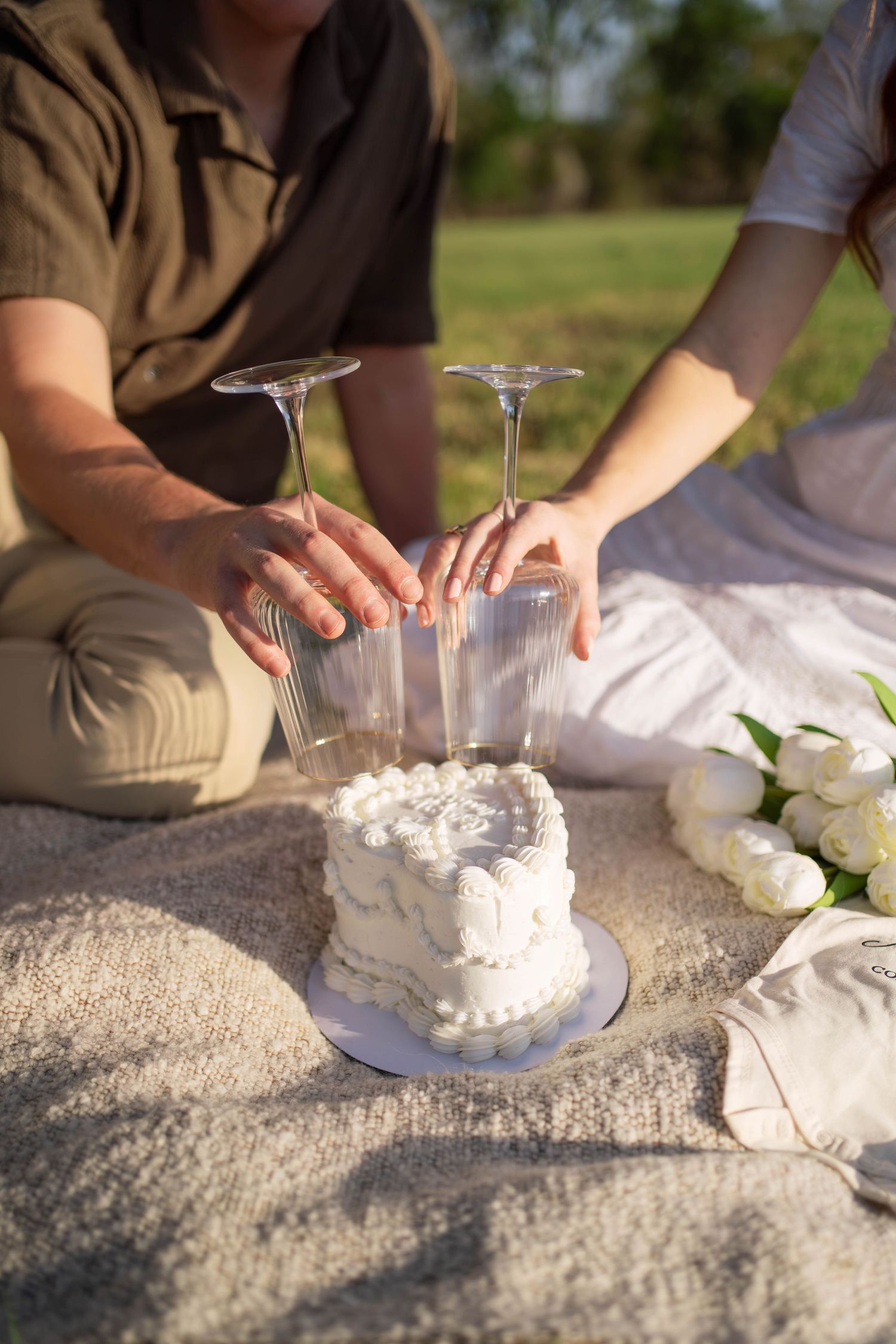 Couple placing wine glasses near a white heart-shaped cake on a picnic blanket.