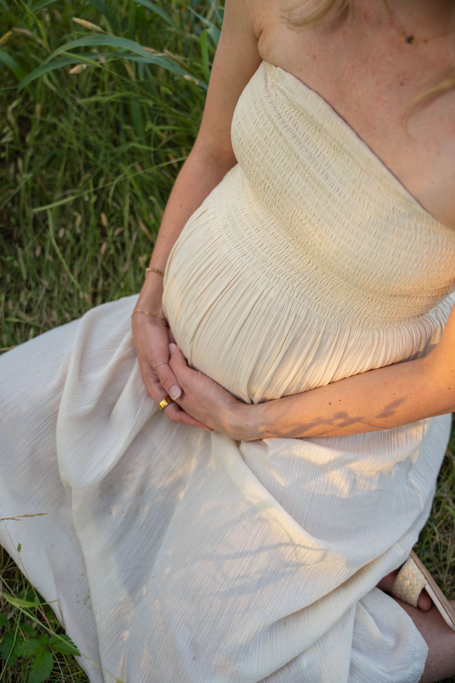 Pregnant woman in cream strapless dress cradling baby bump while sitting in green grass field.