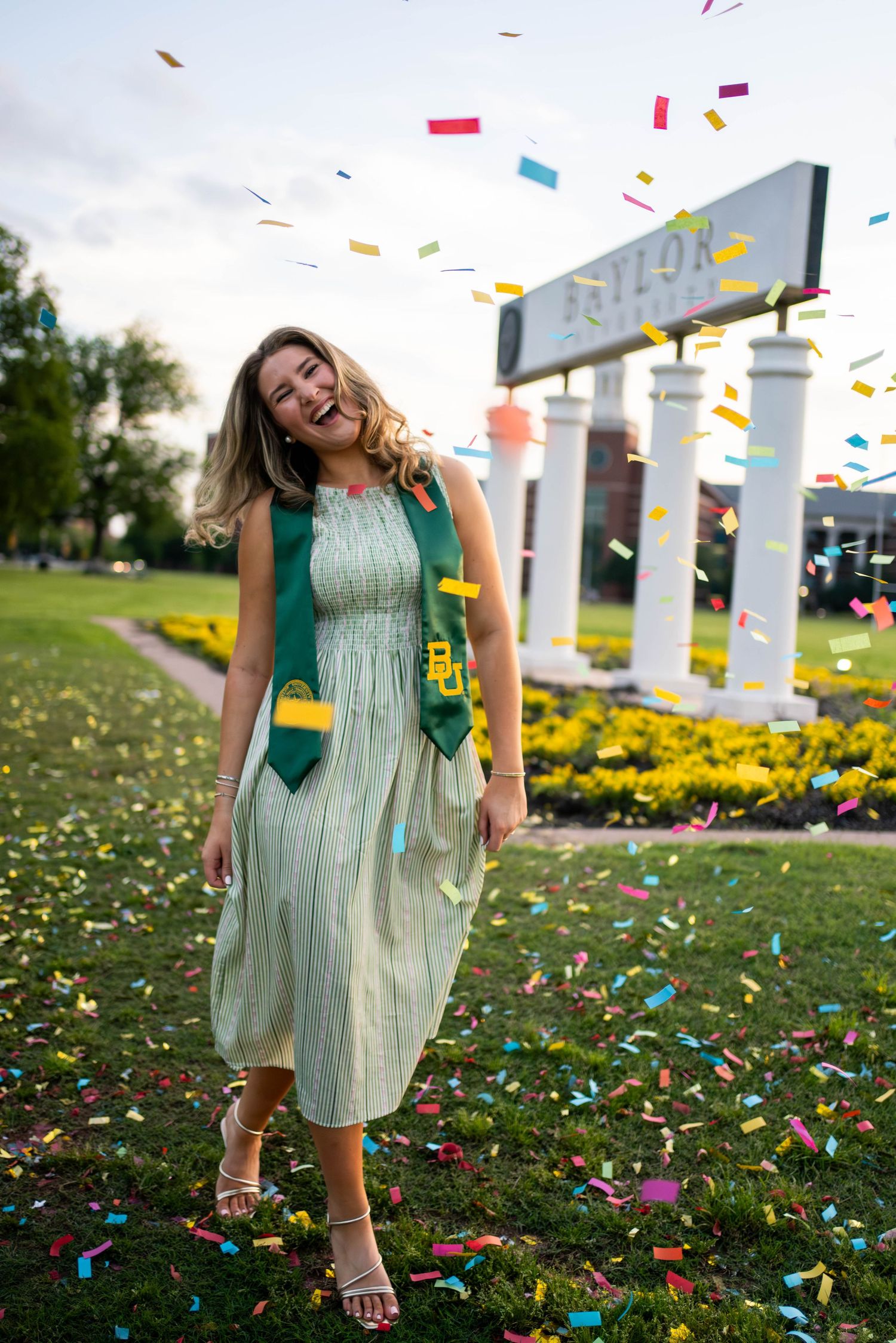 Smiling graduate in green dress and sash celebrates with colorful confetti near Baylor University sign.