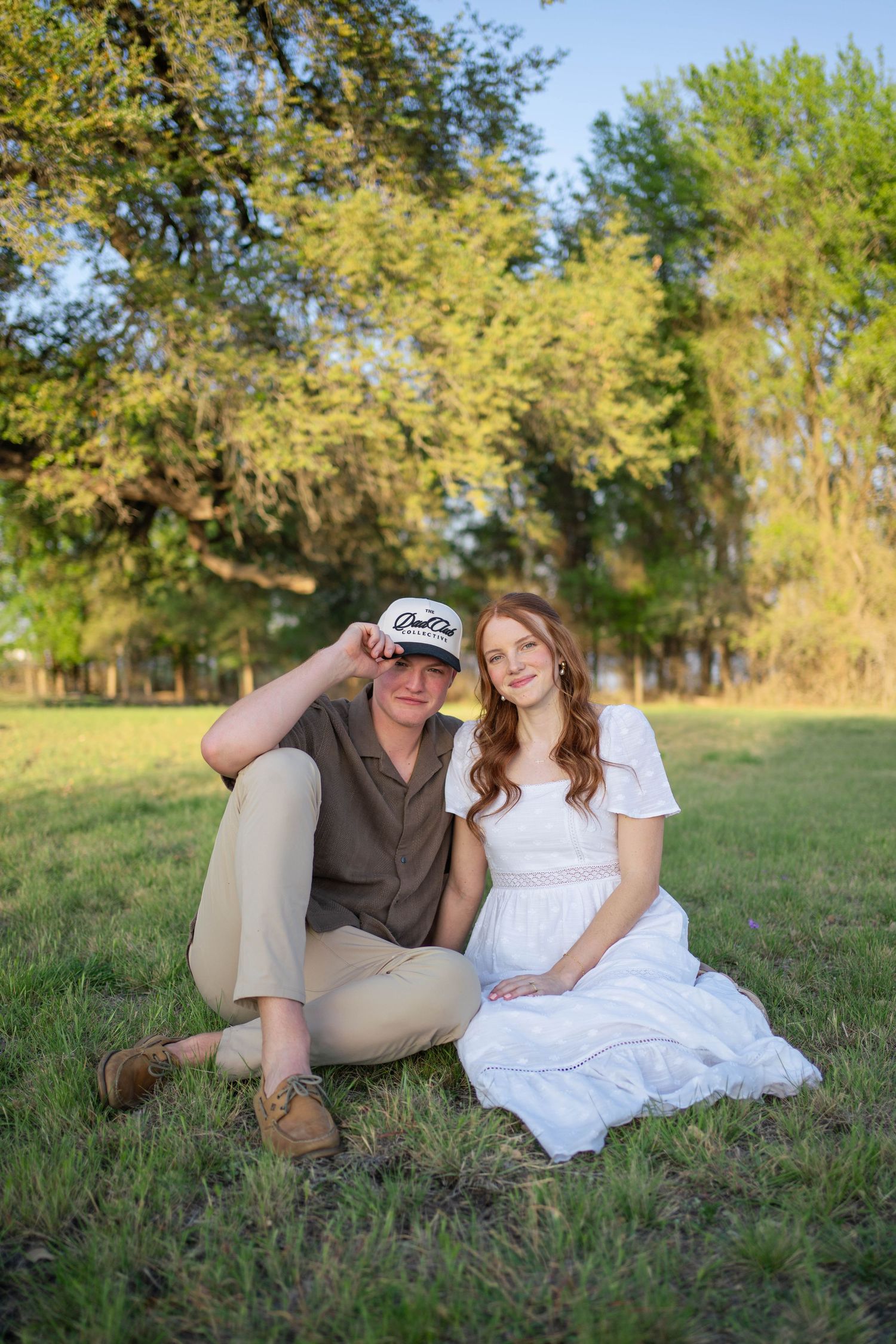 Couple sitting together on green grass under shady trees, woman in white dress touching baby bump.