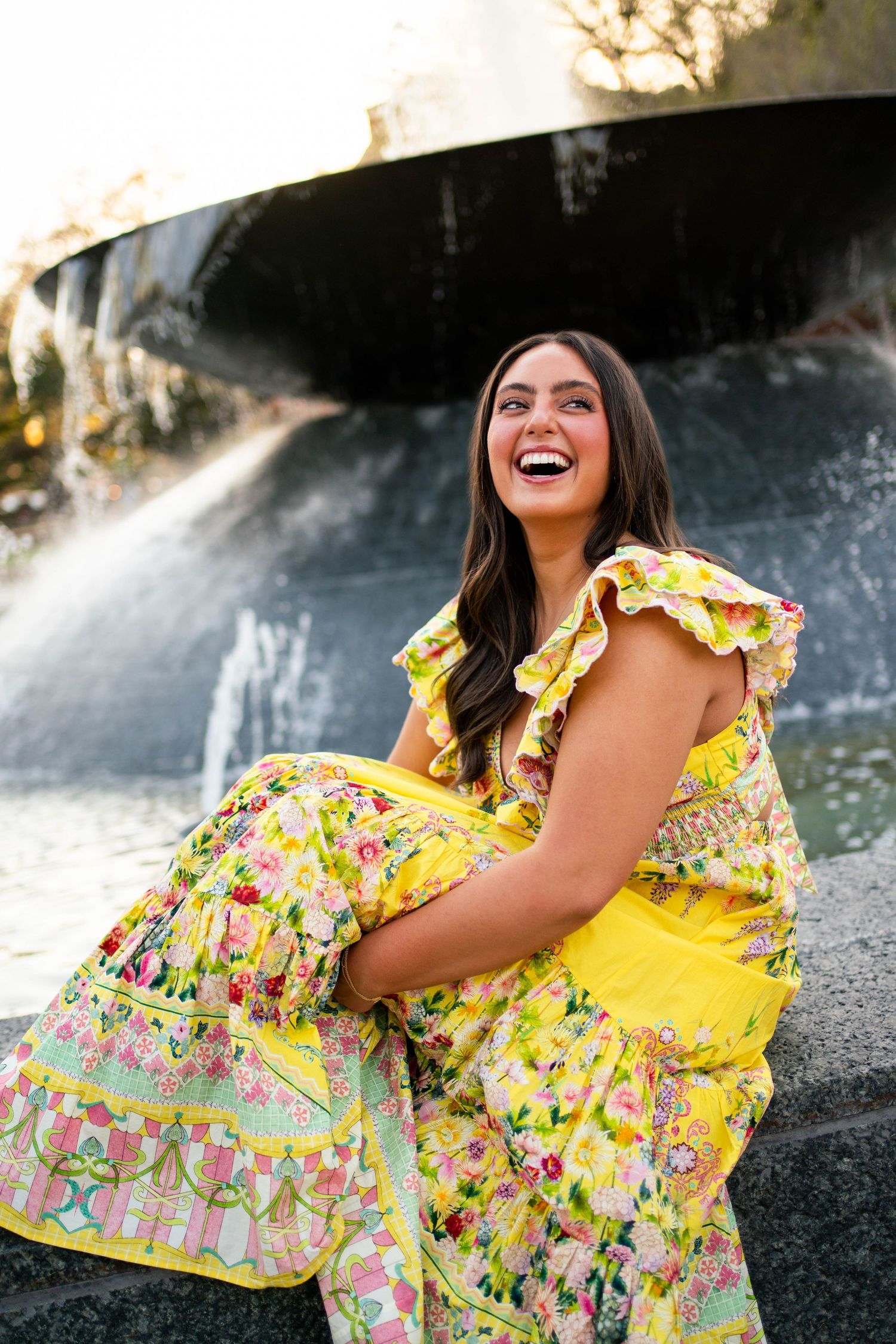 Woman in yellow floral dress laughing joyfully while sitting near a fountain outdoors.