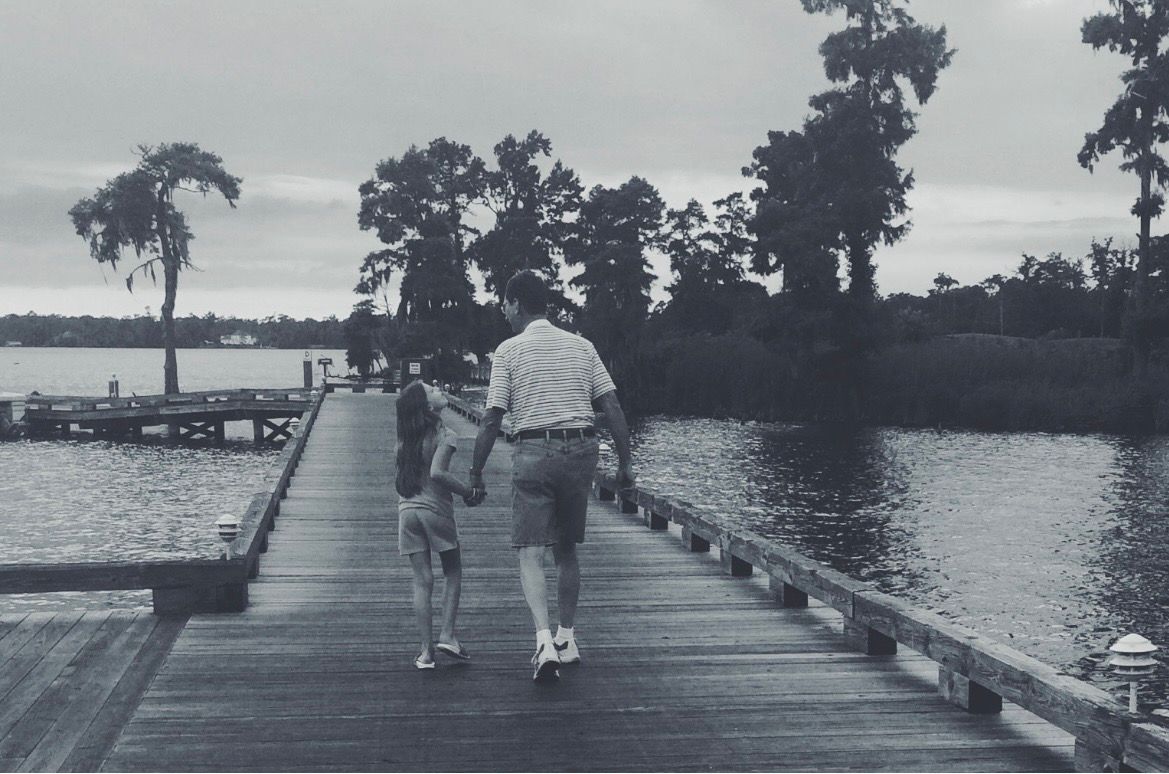 Two people walking together on a wooden dock over calm water surrounded by trees in black and white.