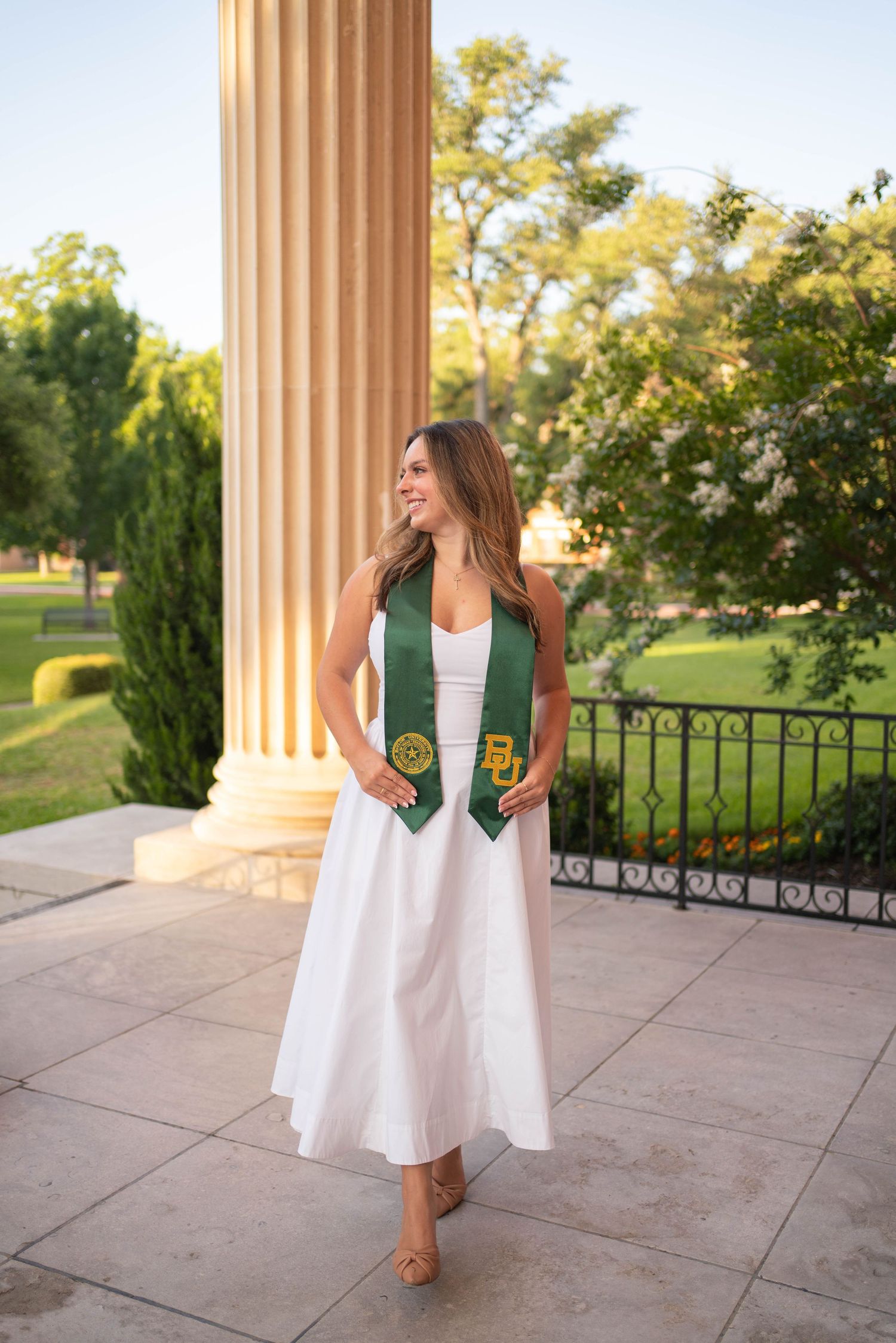 Female Baylor University graduate in white dress with green BU stole posing by large stone column.