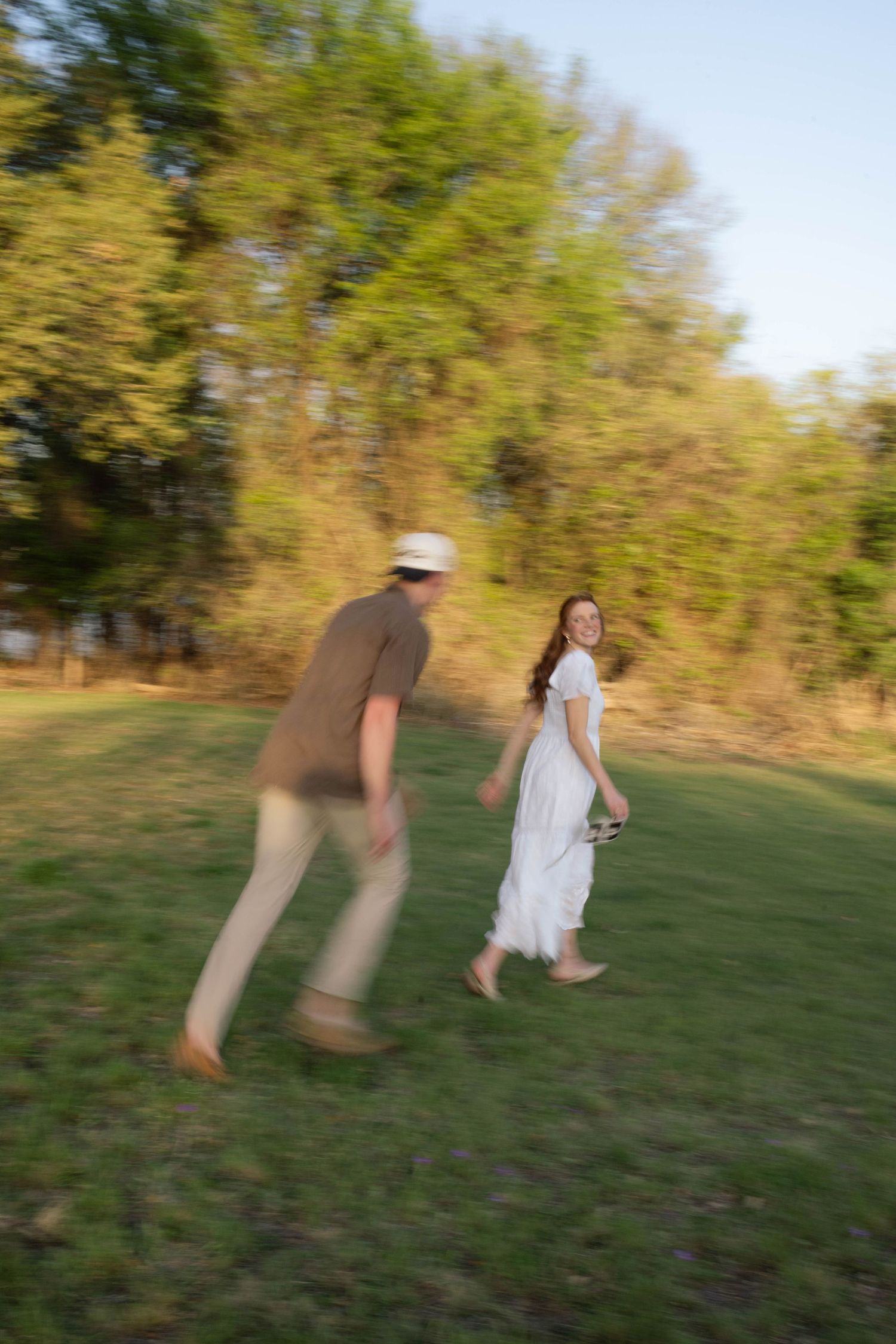 Couple walking in a sunny green meadow, woman in white dress smiling back at man in hat.