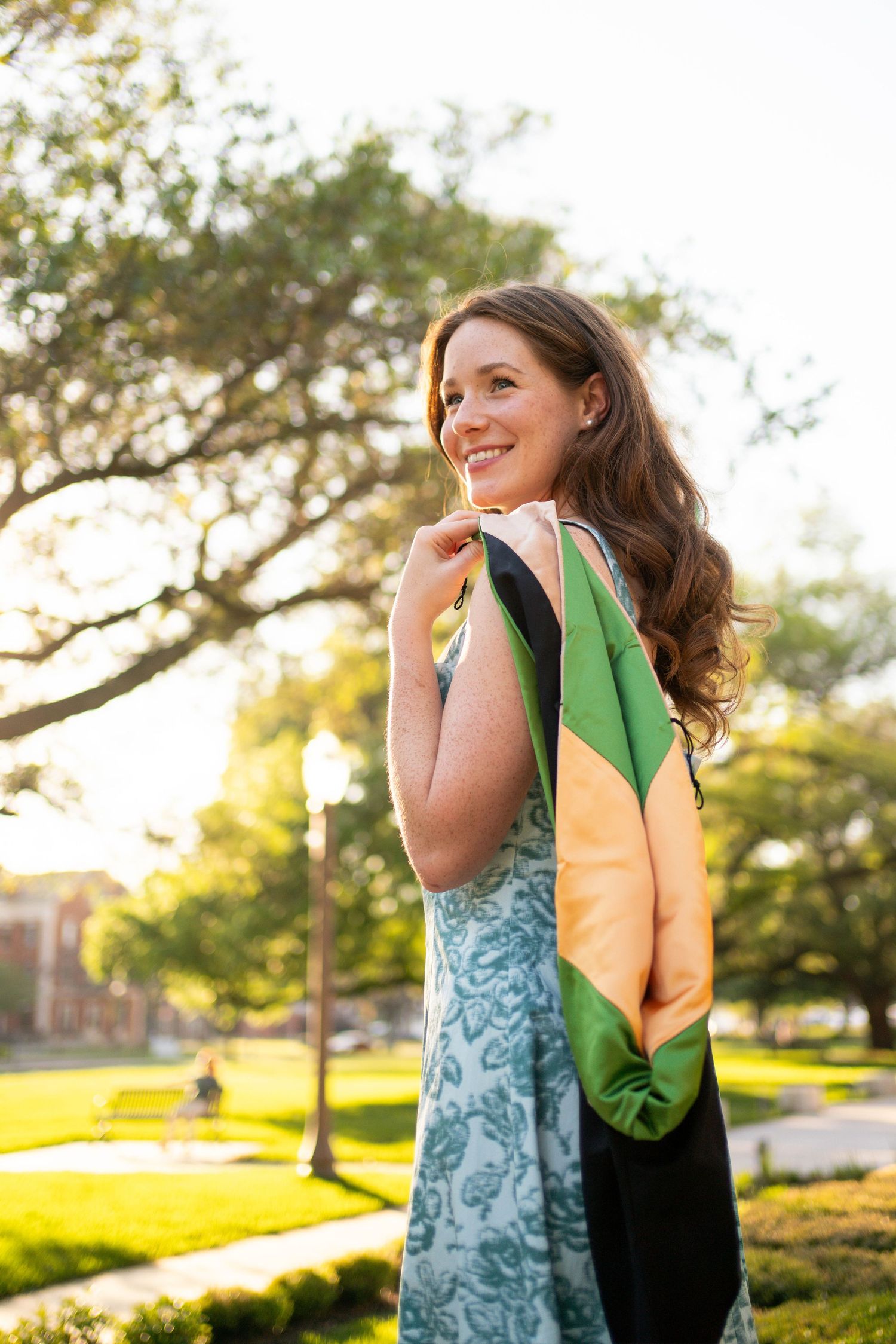 Smiling young woman in floral dress holding green and gold graduation stole in sunny campus park.