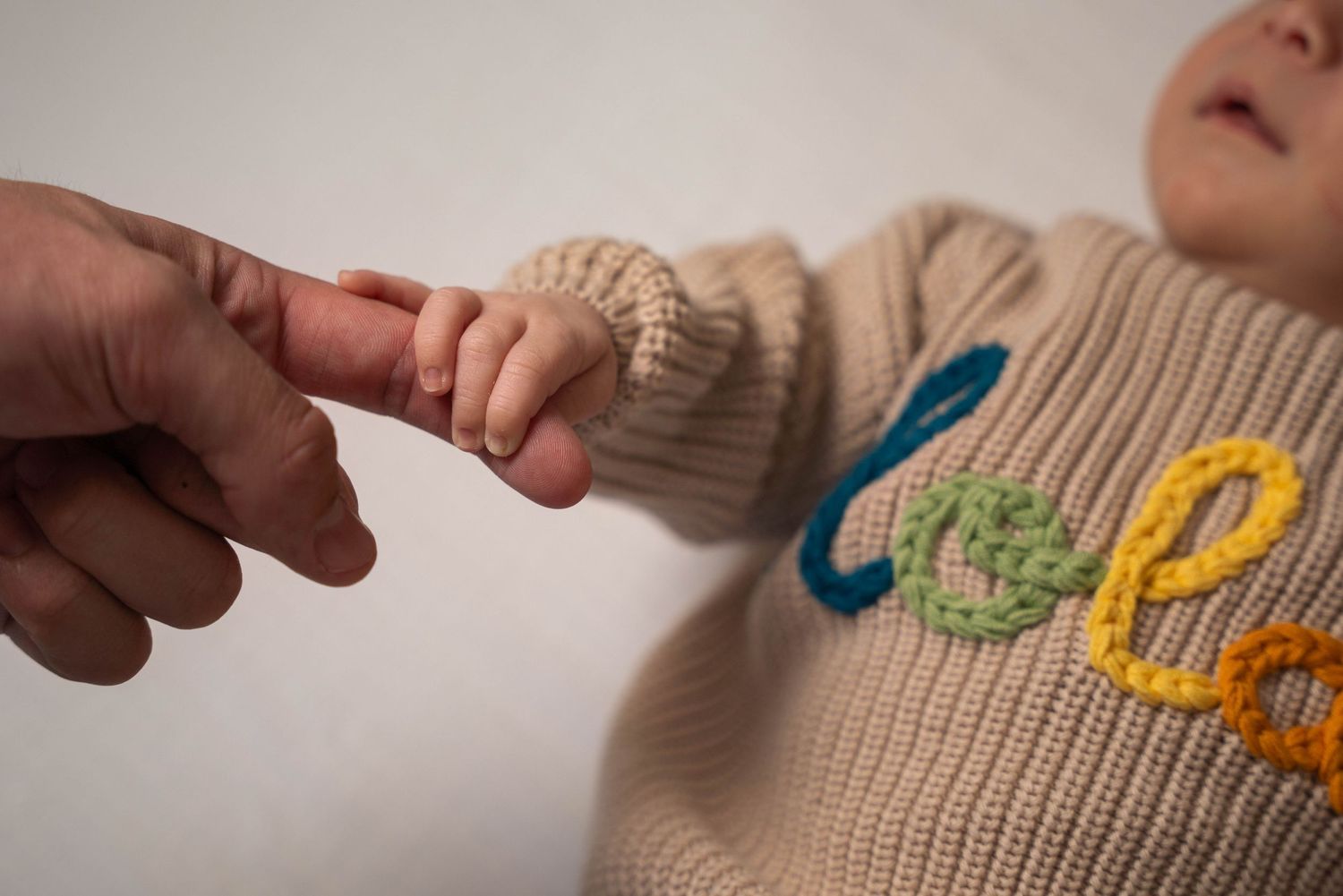 A newborn baby in a beige knit sweater gently grips an adult's finger, symbolizing new life and parental bond.