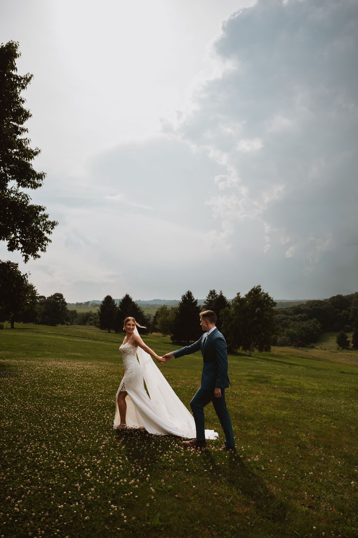 Bride and groom holding hands walking through a lush green meadow under dramatic cloudy skies.