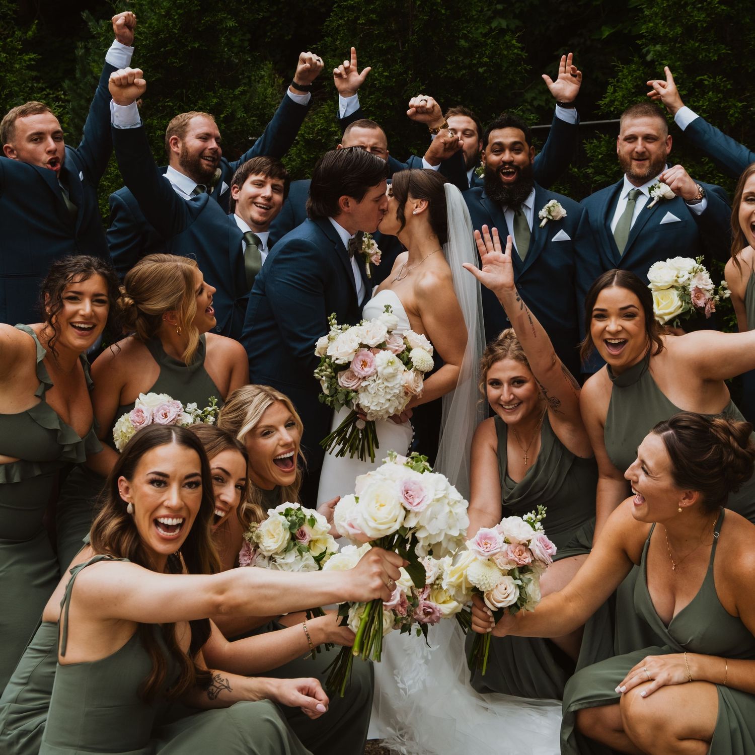 Bride and groom kiss surrounded by cheering wedding party in green bridesmaid dresses and navy suits outdoors.