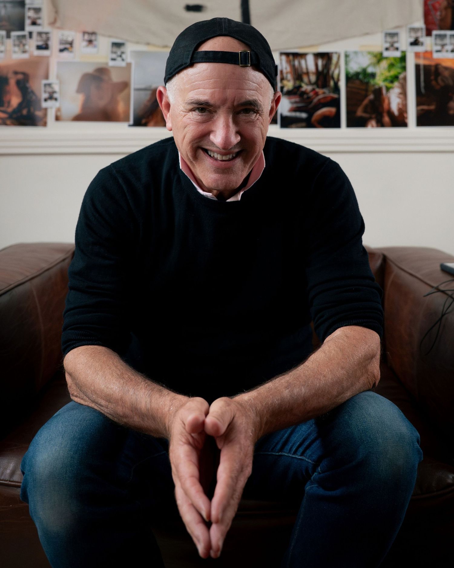 Smiling middle-aged man in black sweater and backwards cap sits relaxed on brown leather couch with photos on wall behind him.