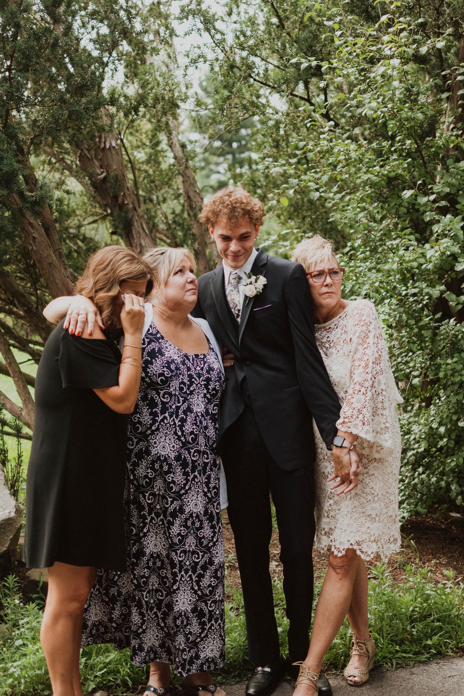 Smiling groom with curly red hair poses with three women in a lush green garden setting.