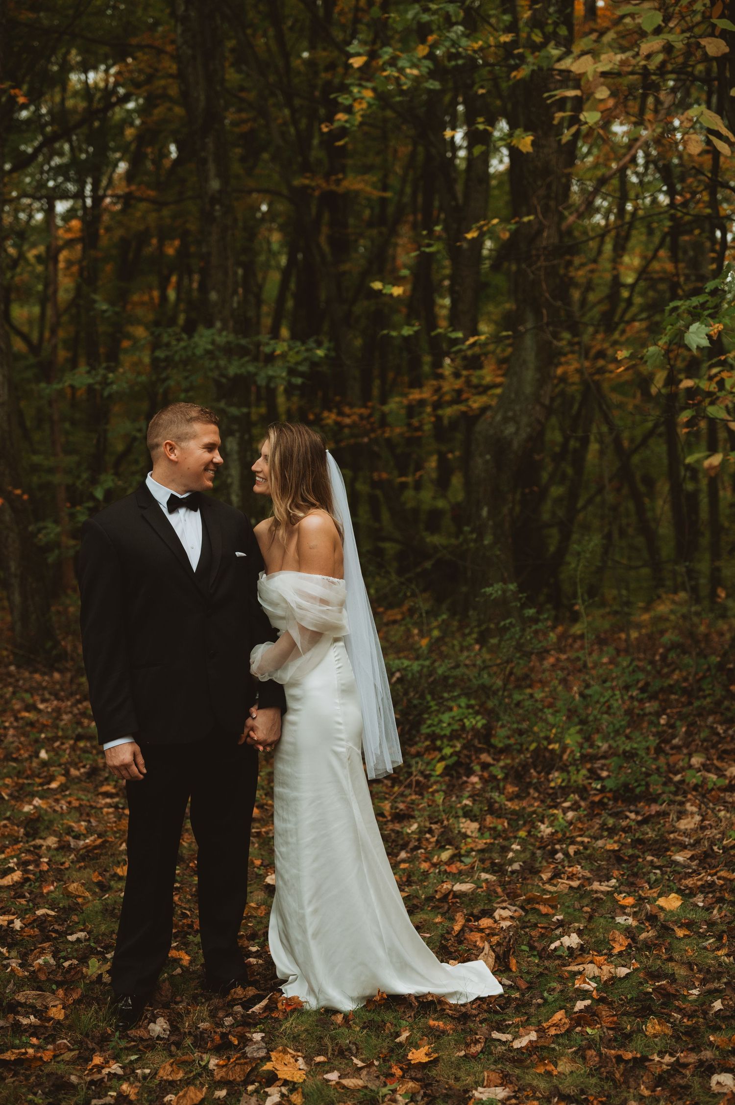 Bride in off-shoulder gown and groom in tuxedo share a smile in a golden autumn forest setting.