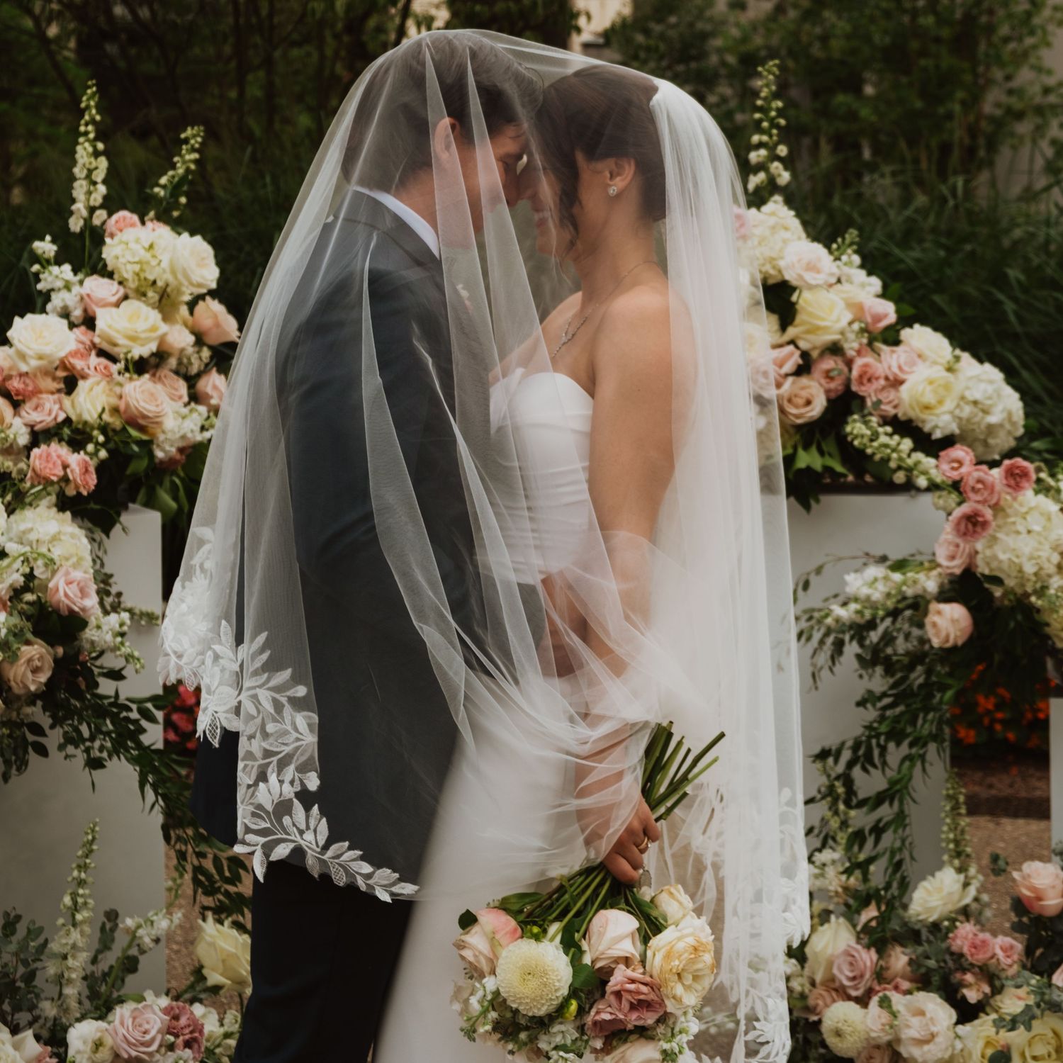 Bride and groom share an intimate moment under a lace veil at a floral ceremony arch with pink roses.