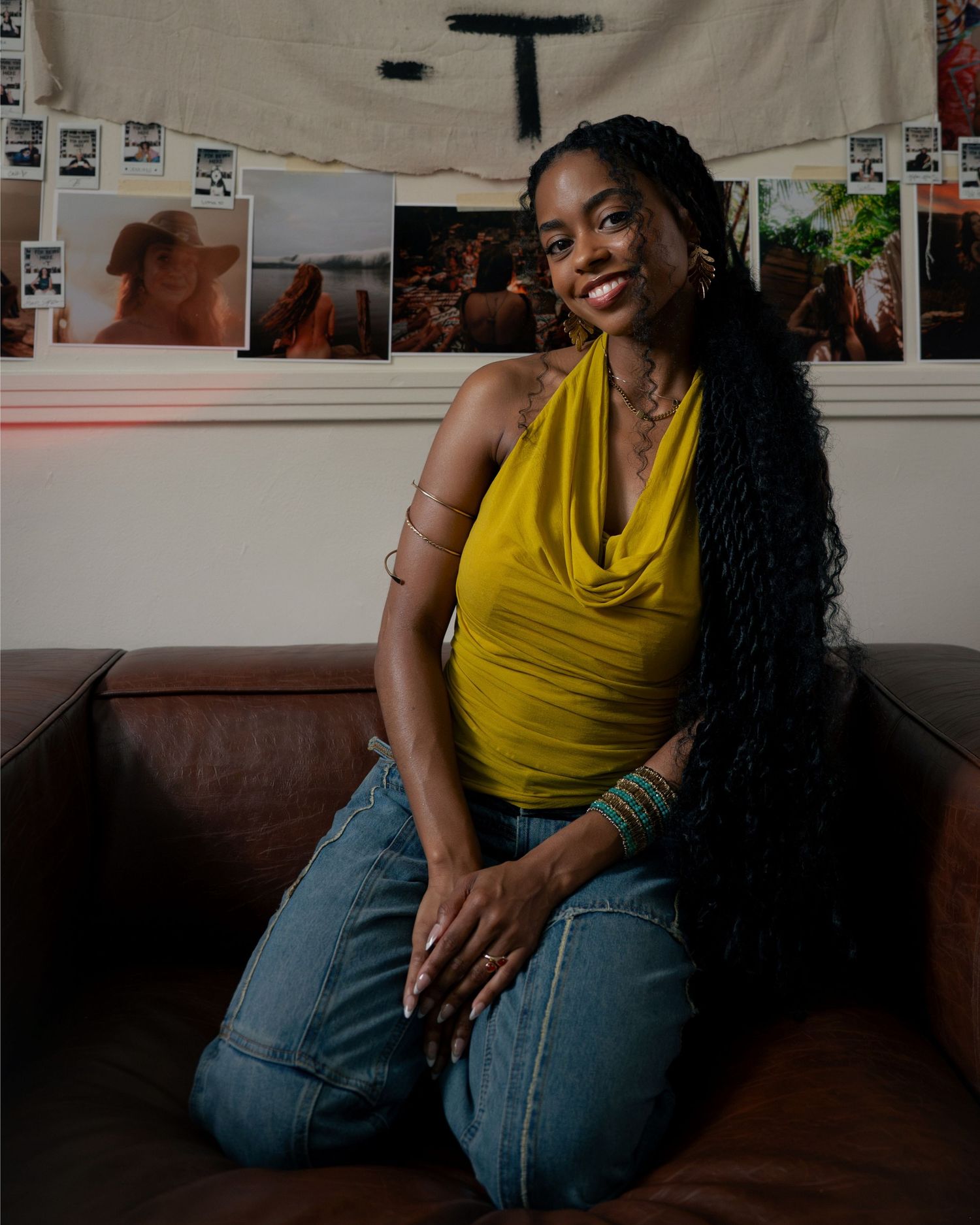 Smiling woman in yellow halter top and jeans sits on leather couch in a warmly lit room.