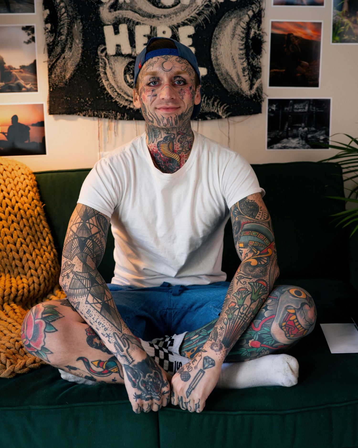 Heavily tattooed man with face and neck tattoos sits cross-legged on green sofa in decorated room.