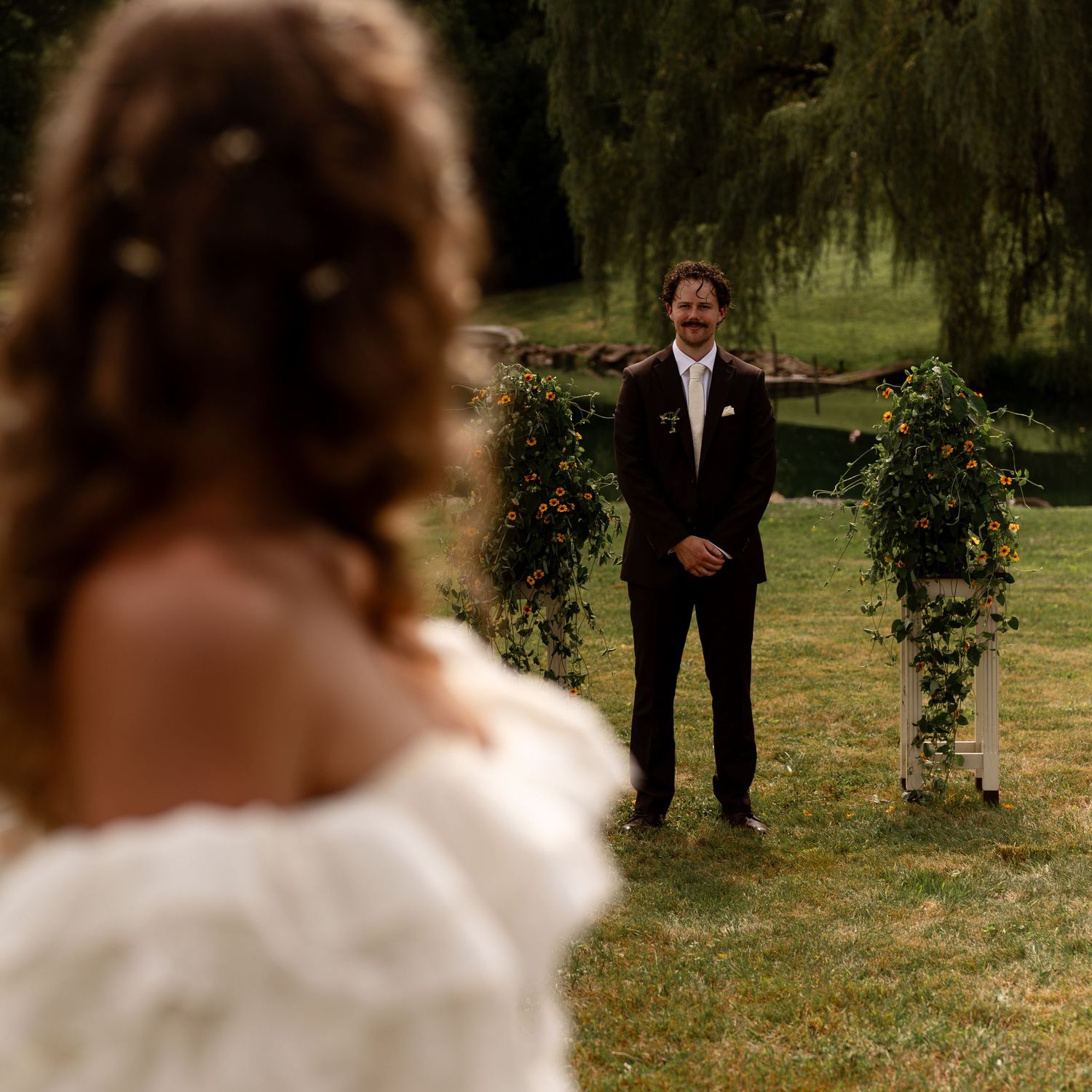 Groom stands at a greenery-adorned outdoor altar smiling as bride approaches across a lawn.