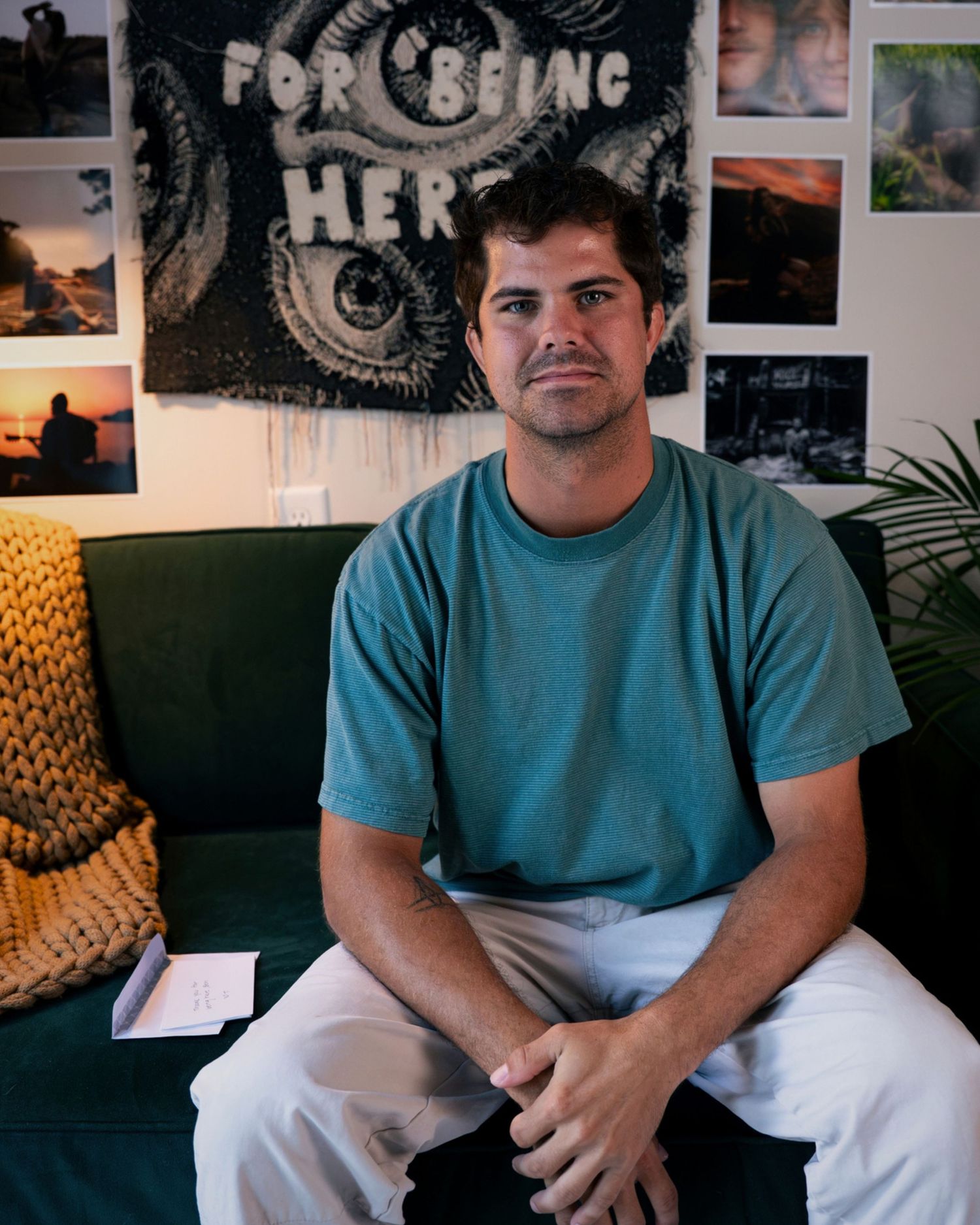 Man in teal shirt sits on green couch with hands clasped in front of a decorative tapestry.
