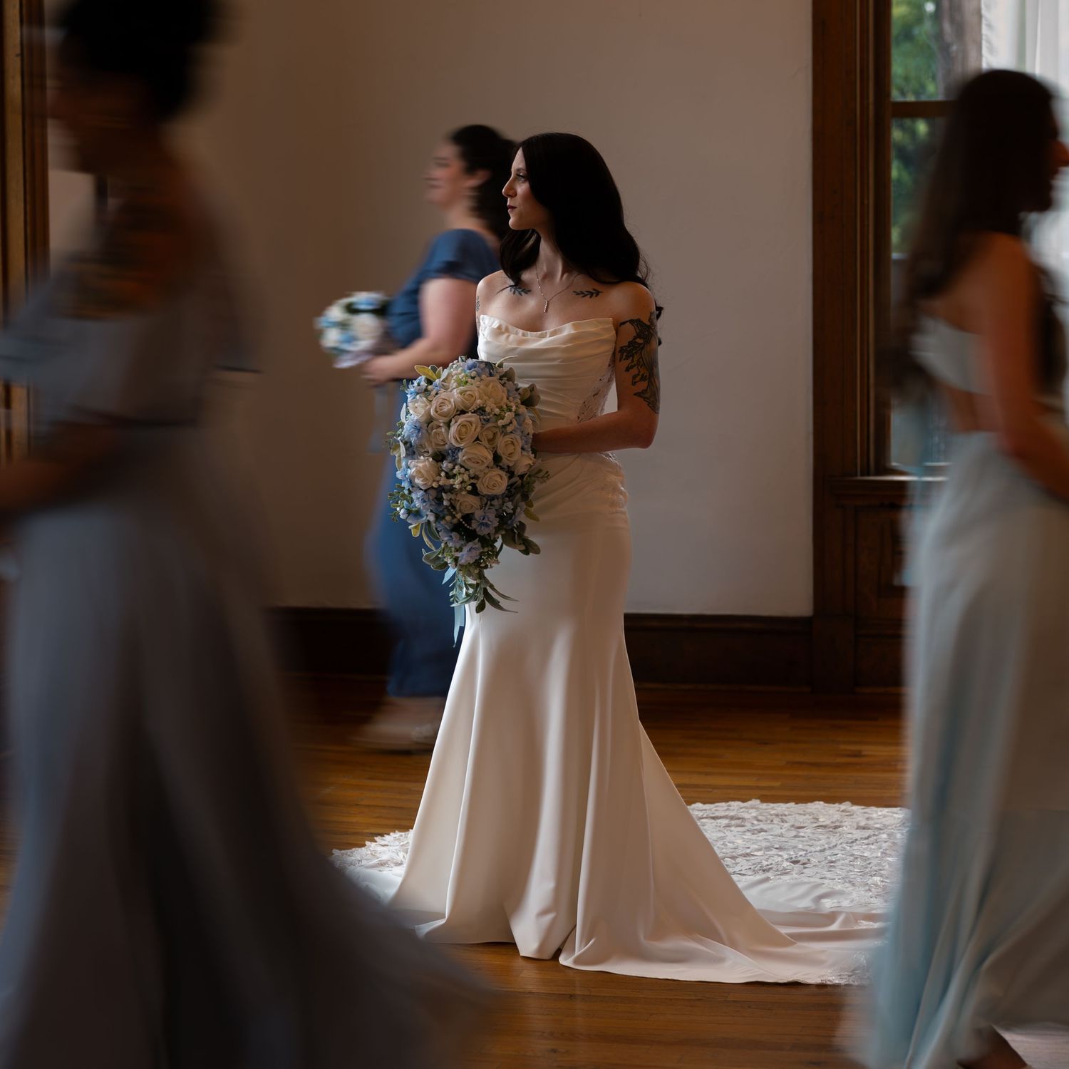 Beautiful dark-haired bride in white gown holds floral bouquet surrounded by bridesmaids.