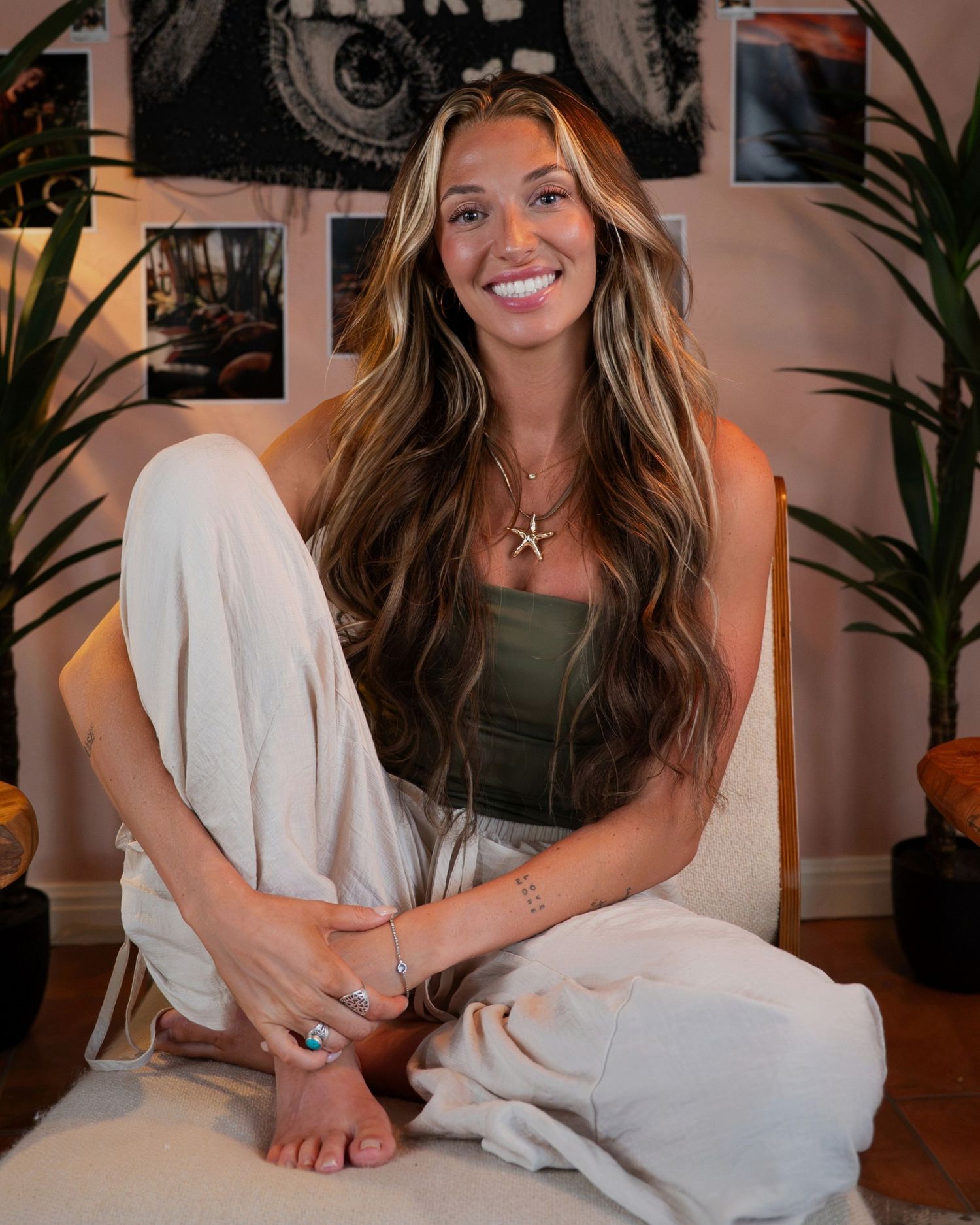 Smiling young woman with highlighted hair sits cross-legged wearing white pants and green top.