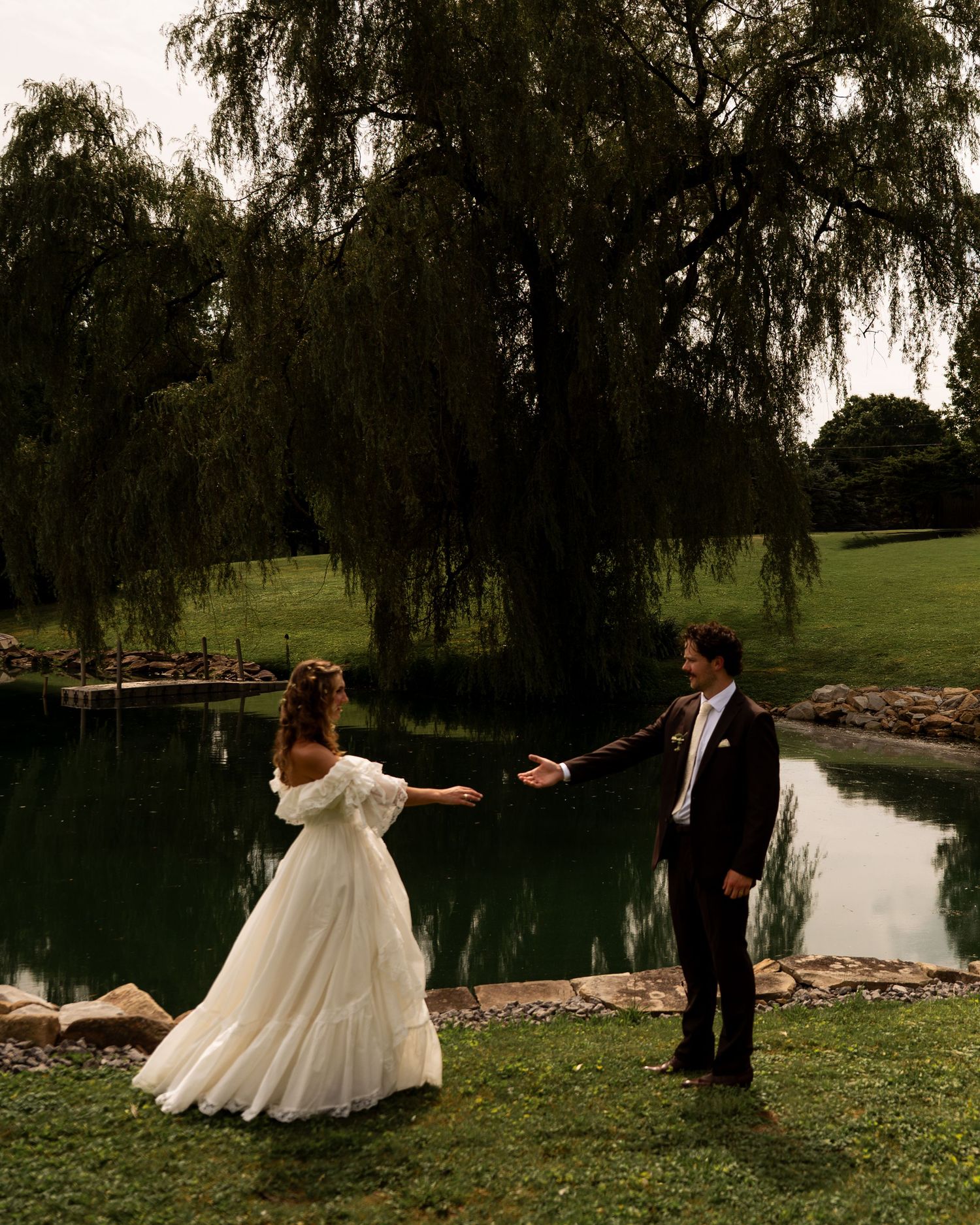 Bride and groom reach for each other near a serene pond beneath a weeping willow tree.