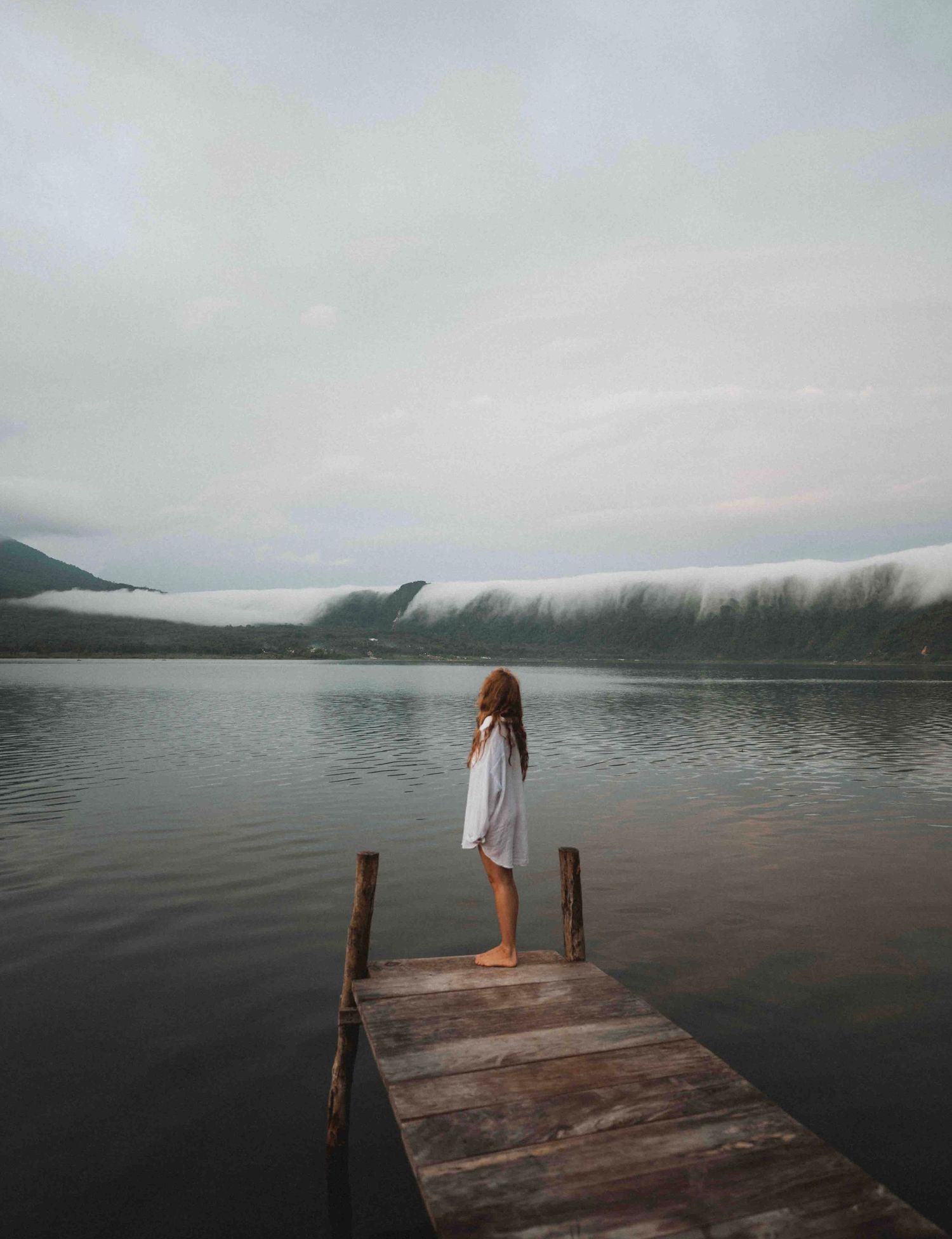 A barefoot girl in a white dress stands on a wooden dock overlooking a misty mountain lake under cloudy skies.