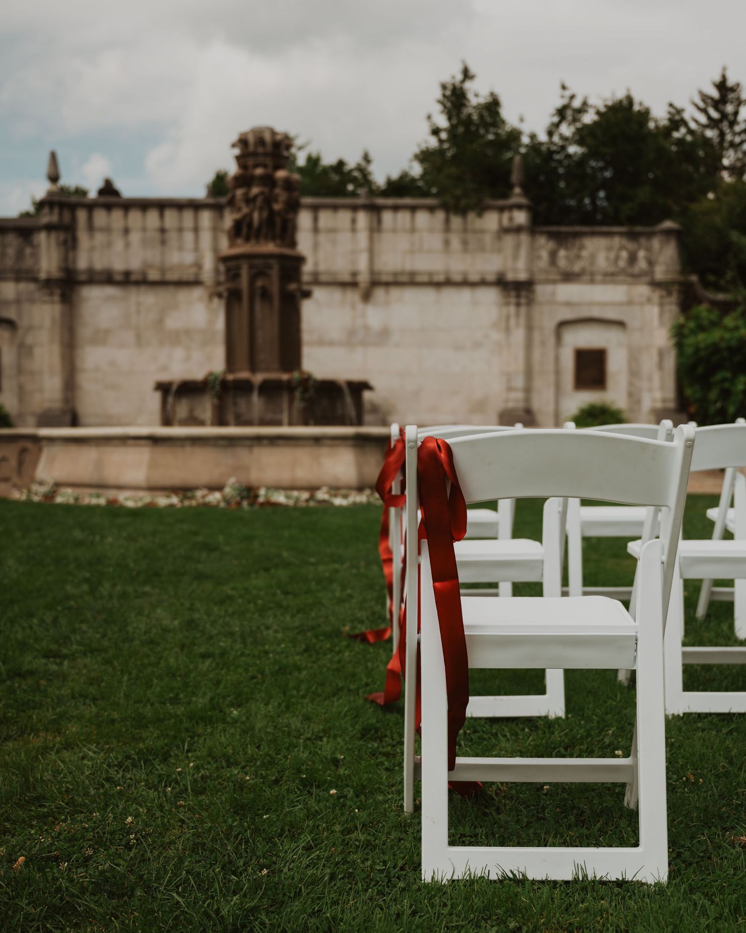 White folding chairs with red ribbon bows set up on lawn before stone wedding venue.