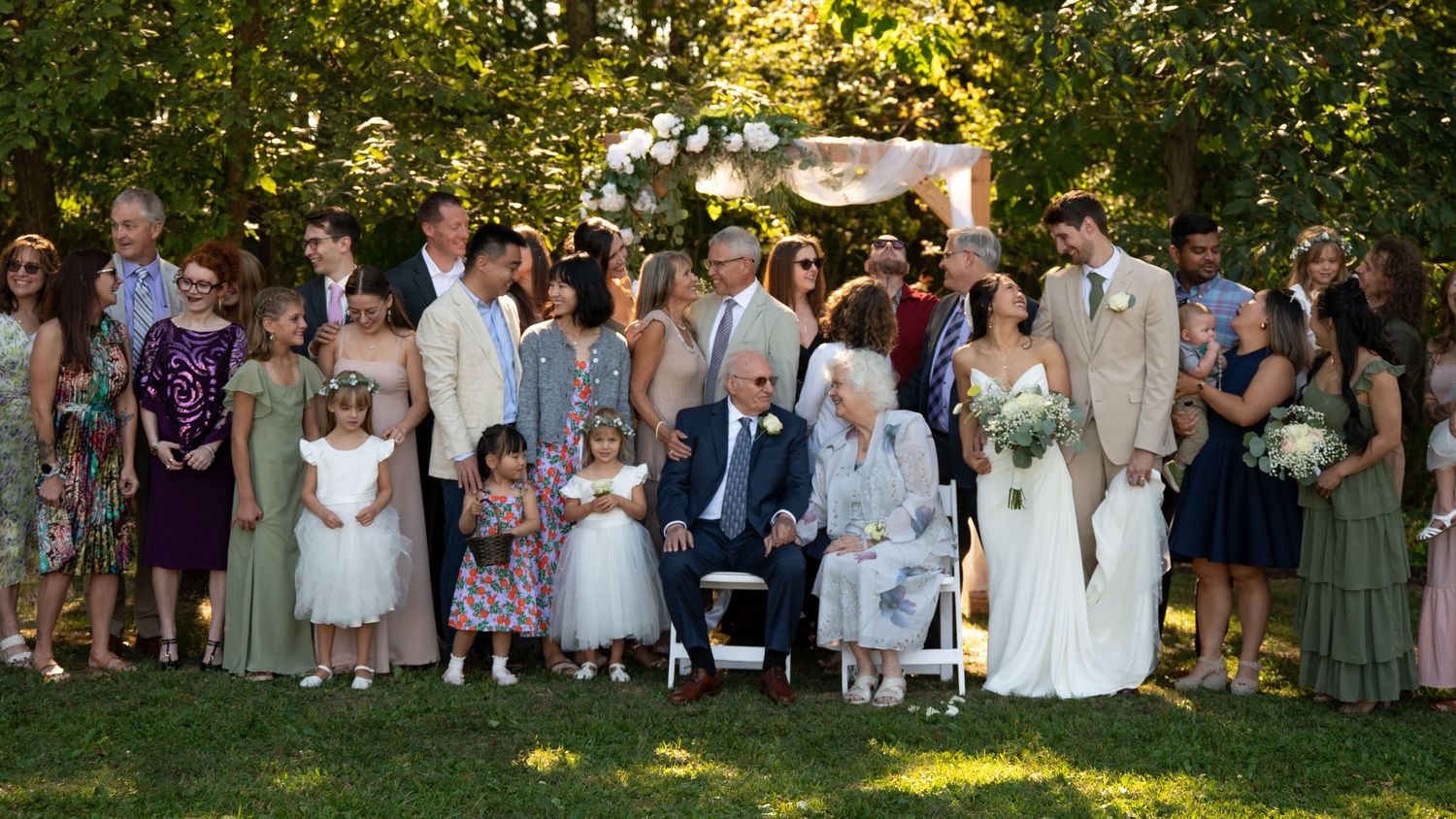 Large wedding family portrait gathered outdoors under a floral arch in a garden setting.