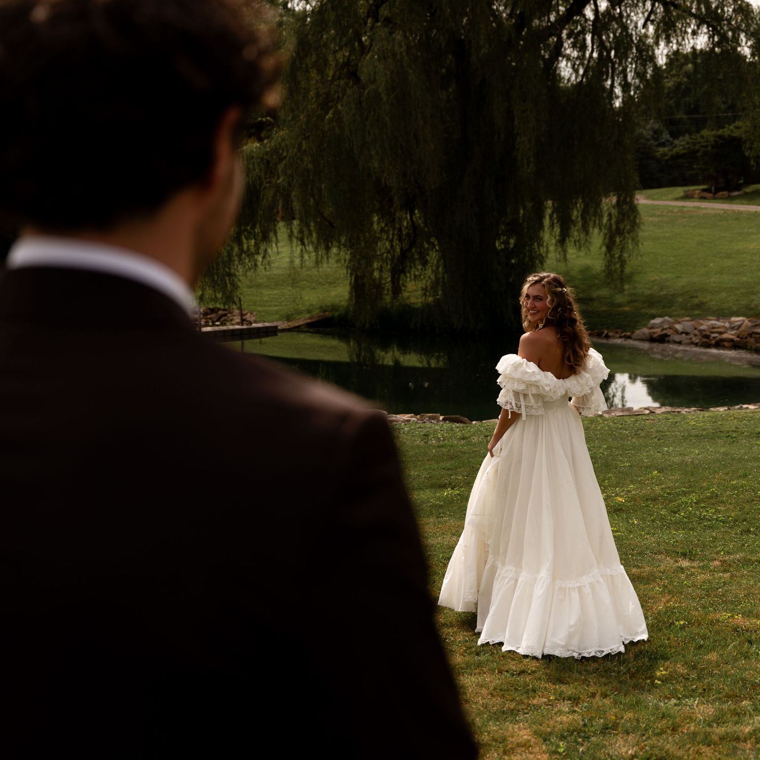 Bride in a white off-shoulder gown stands near a pond under a weeping willow tree.