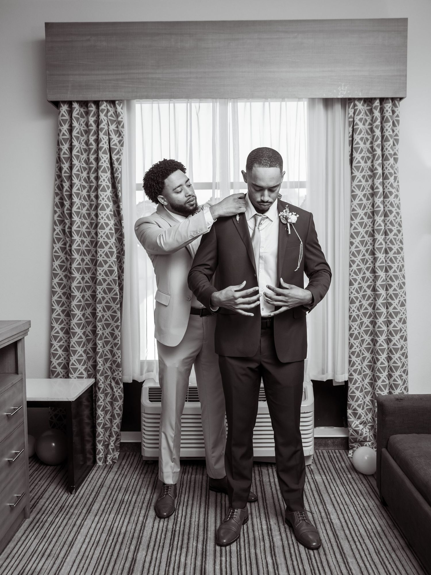 Black-and-white wedding photograph of the groom getting help adjusting his jacket.