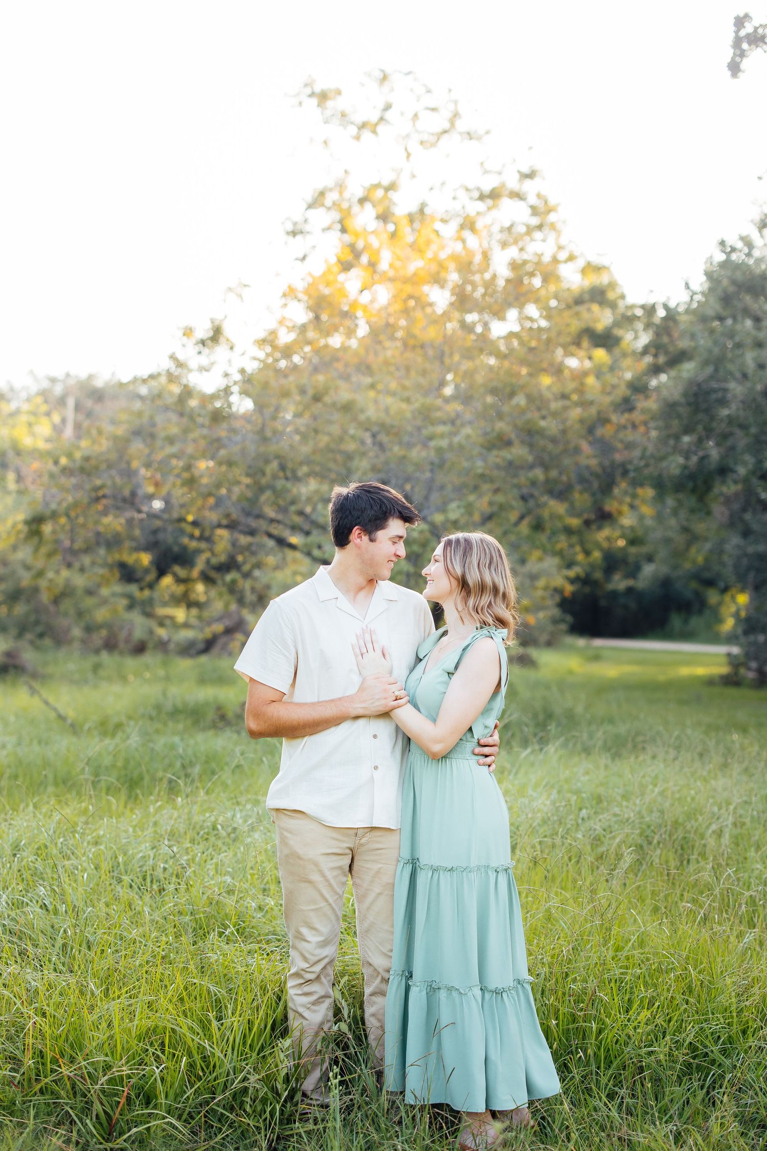 A couple shares a romantic moment in a sunlit meadow, with her wearing a flowing mint green dress and him in casual attire.
