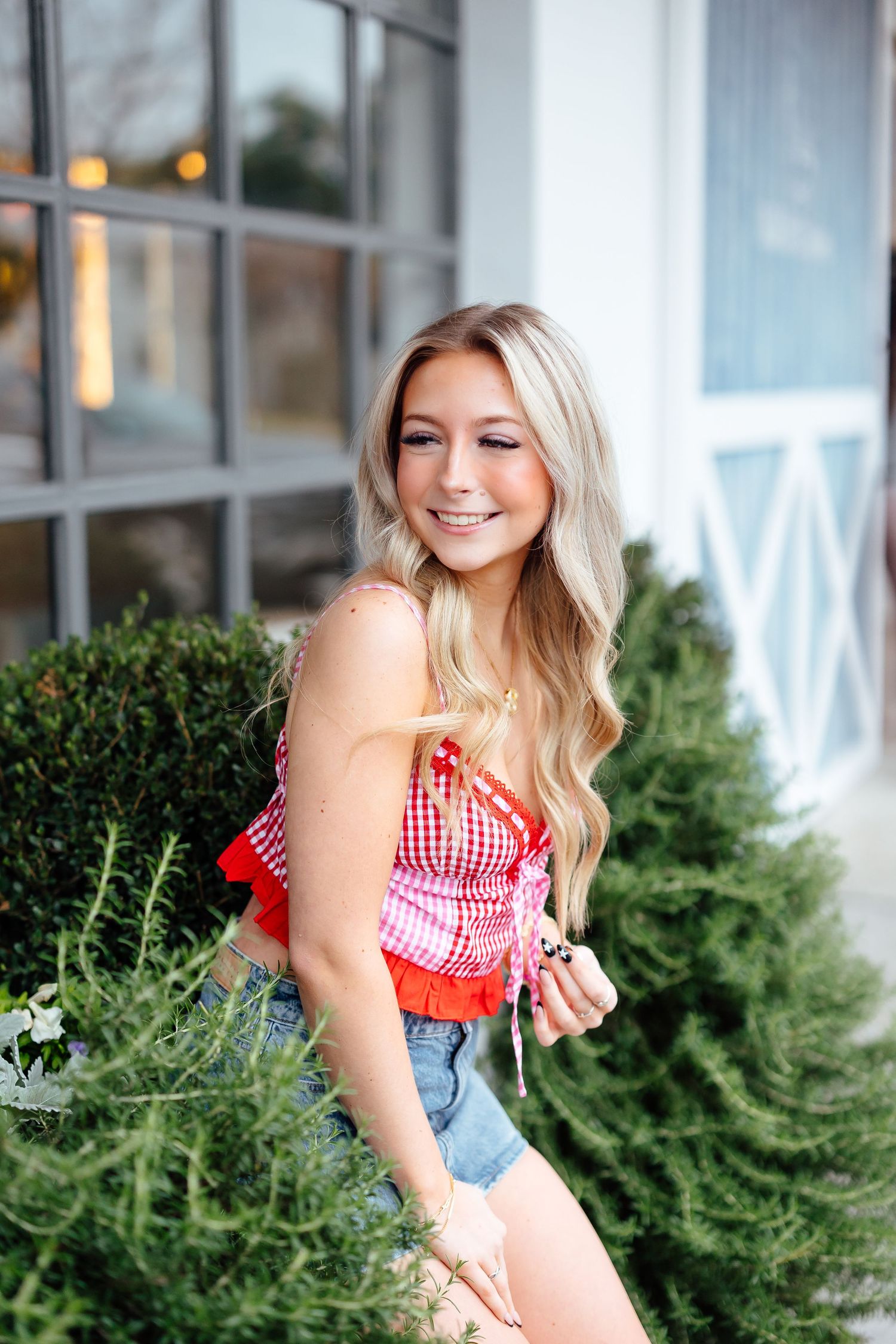 A high school senior in a red gingham crop top and denim shorts poses cheerfully outdoors near houston greenery and a window.