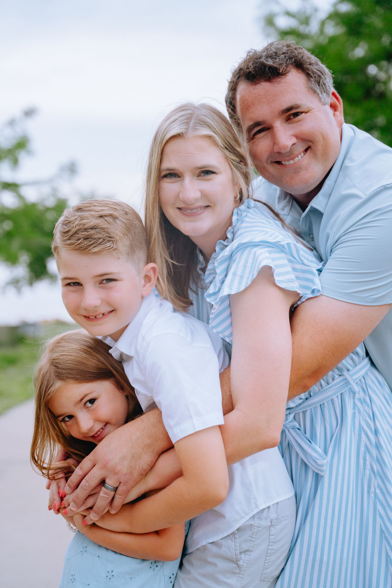 Happy family of four hugging outdoors, parents with two children dressed in coordinating blue and white outfits.