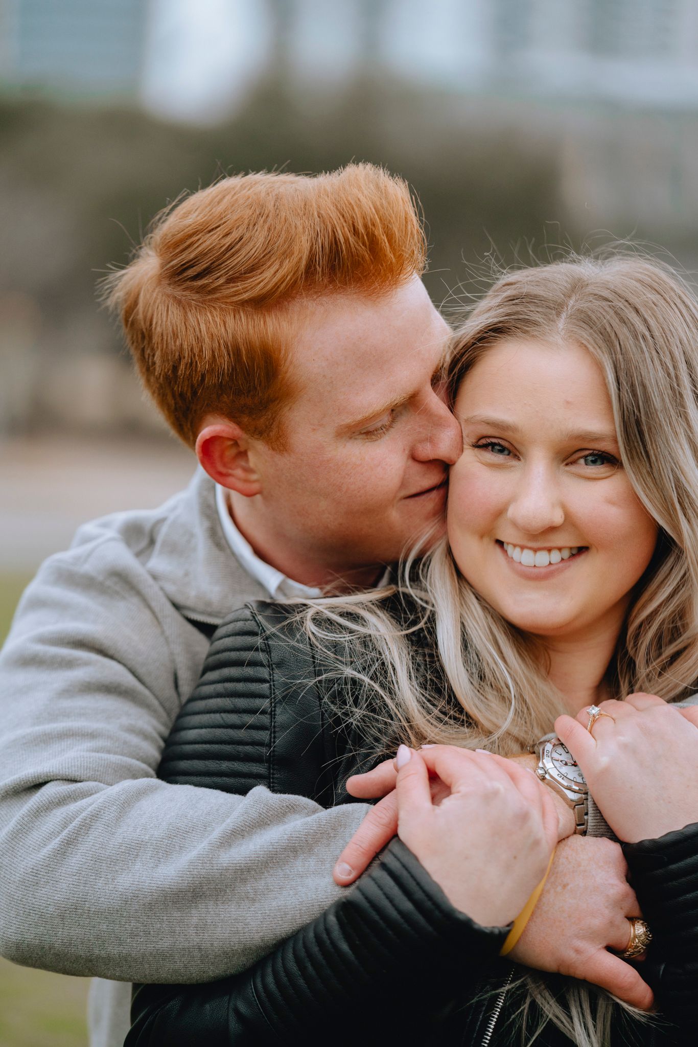 Red-haired man kissing smiling blonde woman on cheek outdoors.
