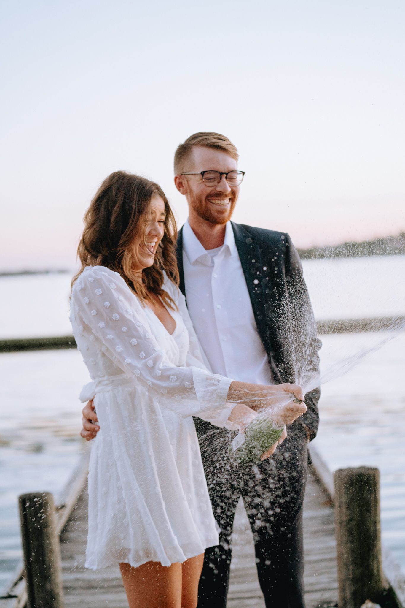 Happy couple popping champagne bottle on wooden dock at sunset, woman in white dress and man in navy blazer.