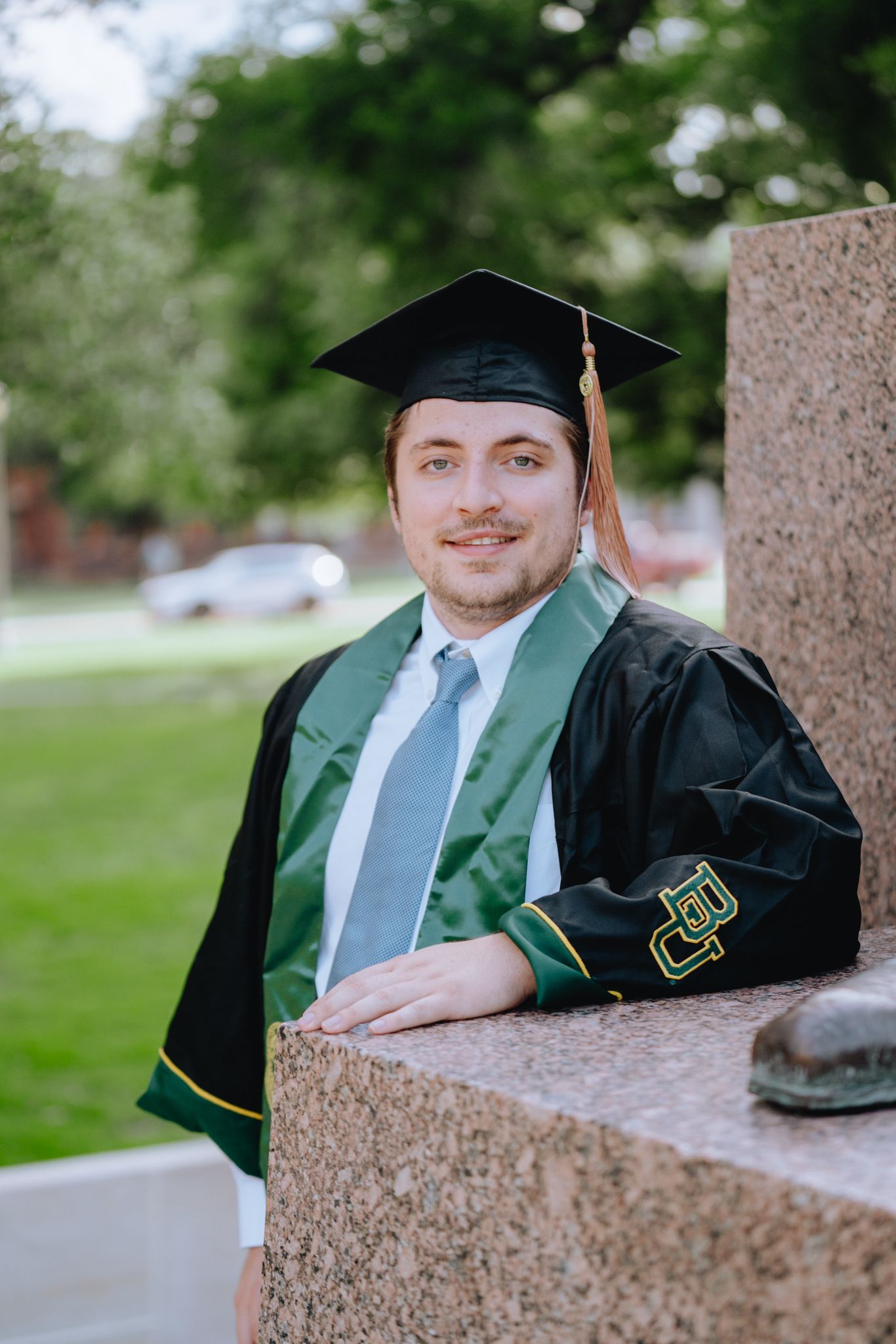 Male Baylor University graduate in black cap and gown with green stole leans against stone pillar.