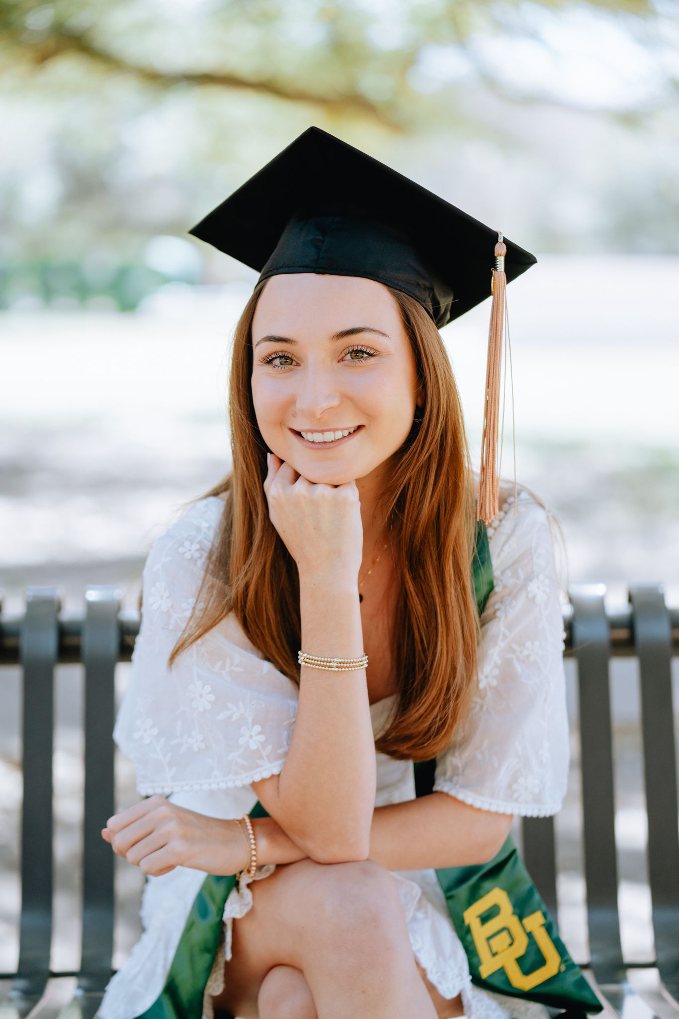 Smiling graduate in cap wearing BU stole seated on bench resting chin on hand.