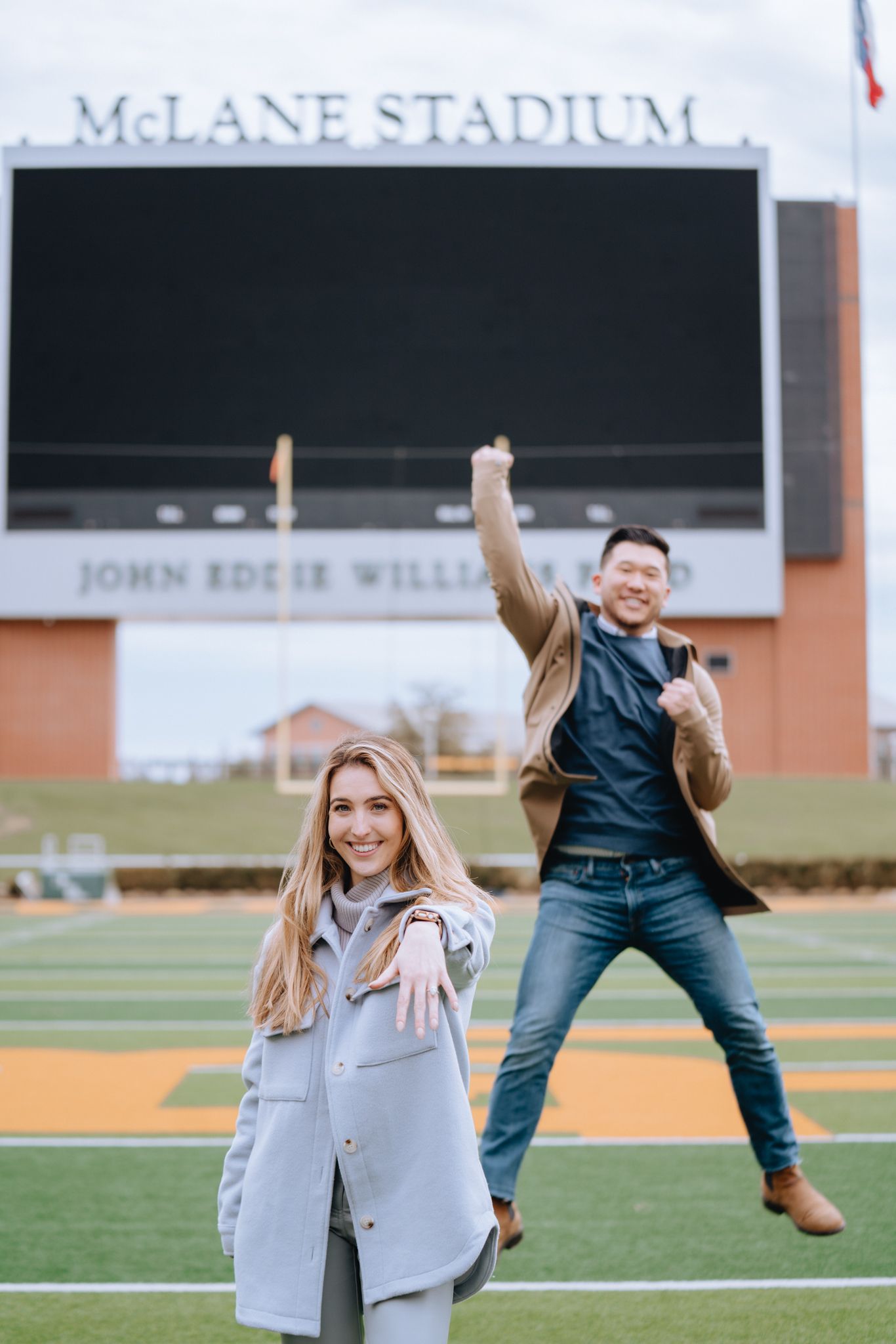 Excited couple celebrating engagement at McLane Stadium, woman showing ring.