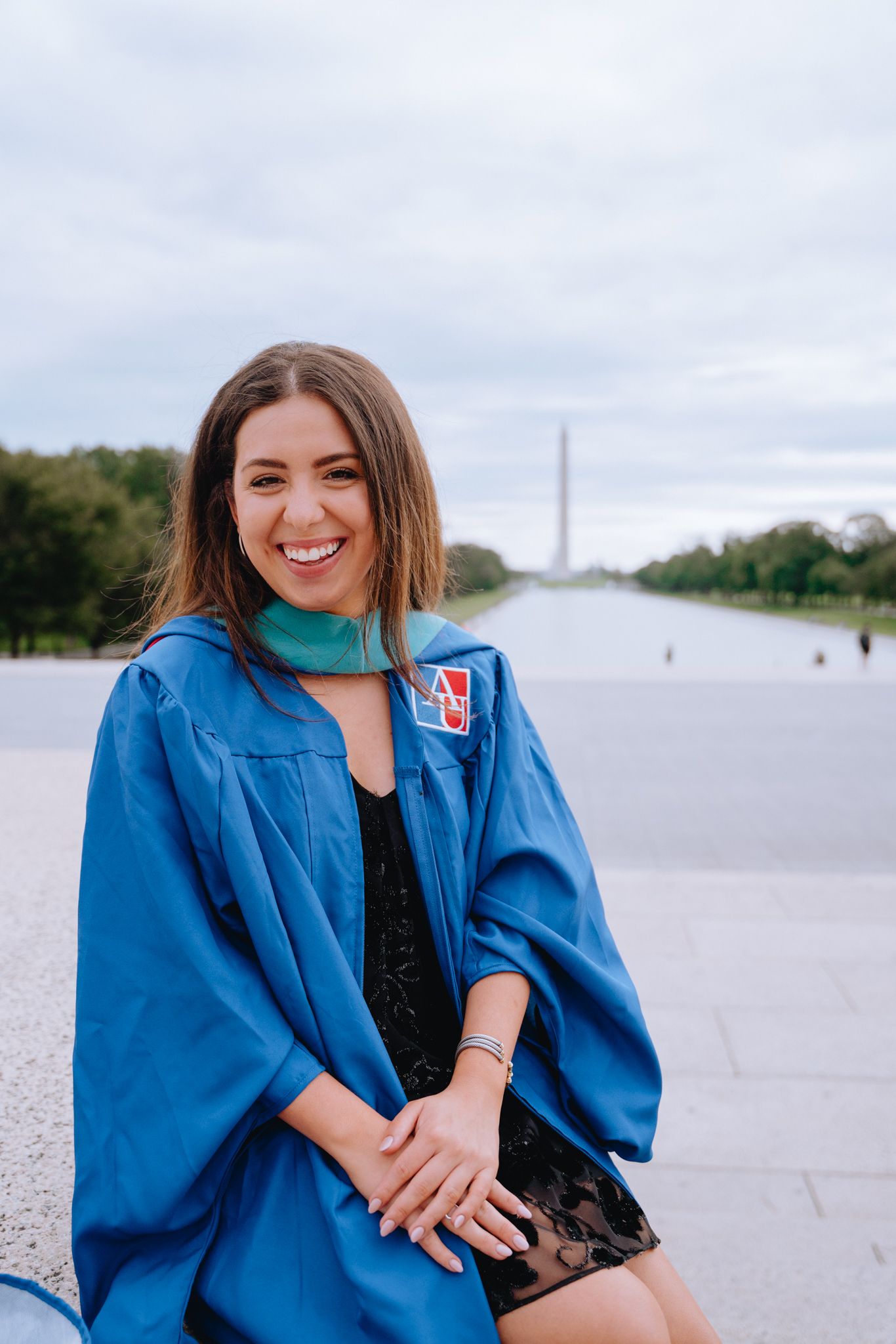 Smiling female graduate in blue cap and gown sits near Washington Monument reflecting pool.