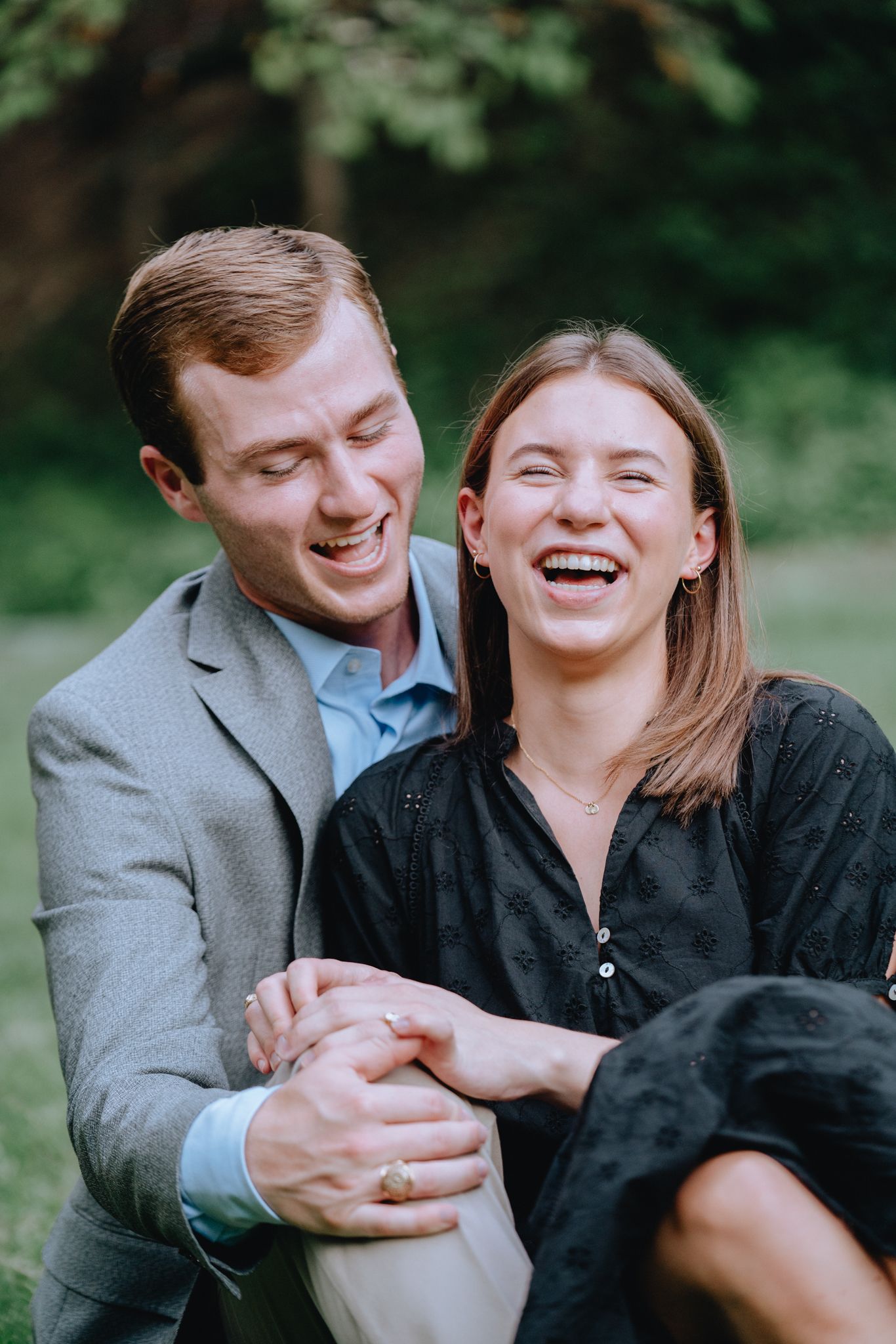Laughing couple sitting outdoors in green park setting, man in gray blazer embracing smiling woman.