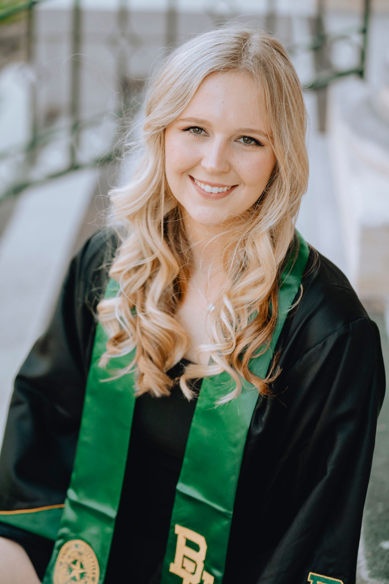 Smiling blonde woman in Baylor University black graduation gown with green stole seated outdoors.