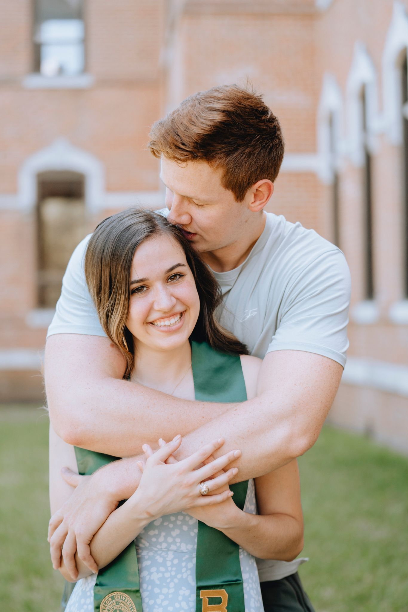 Smiling Baylor University graduate in green stole embraced by partner in front of brick building.