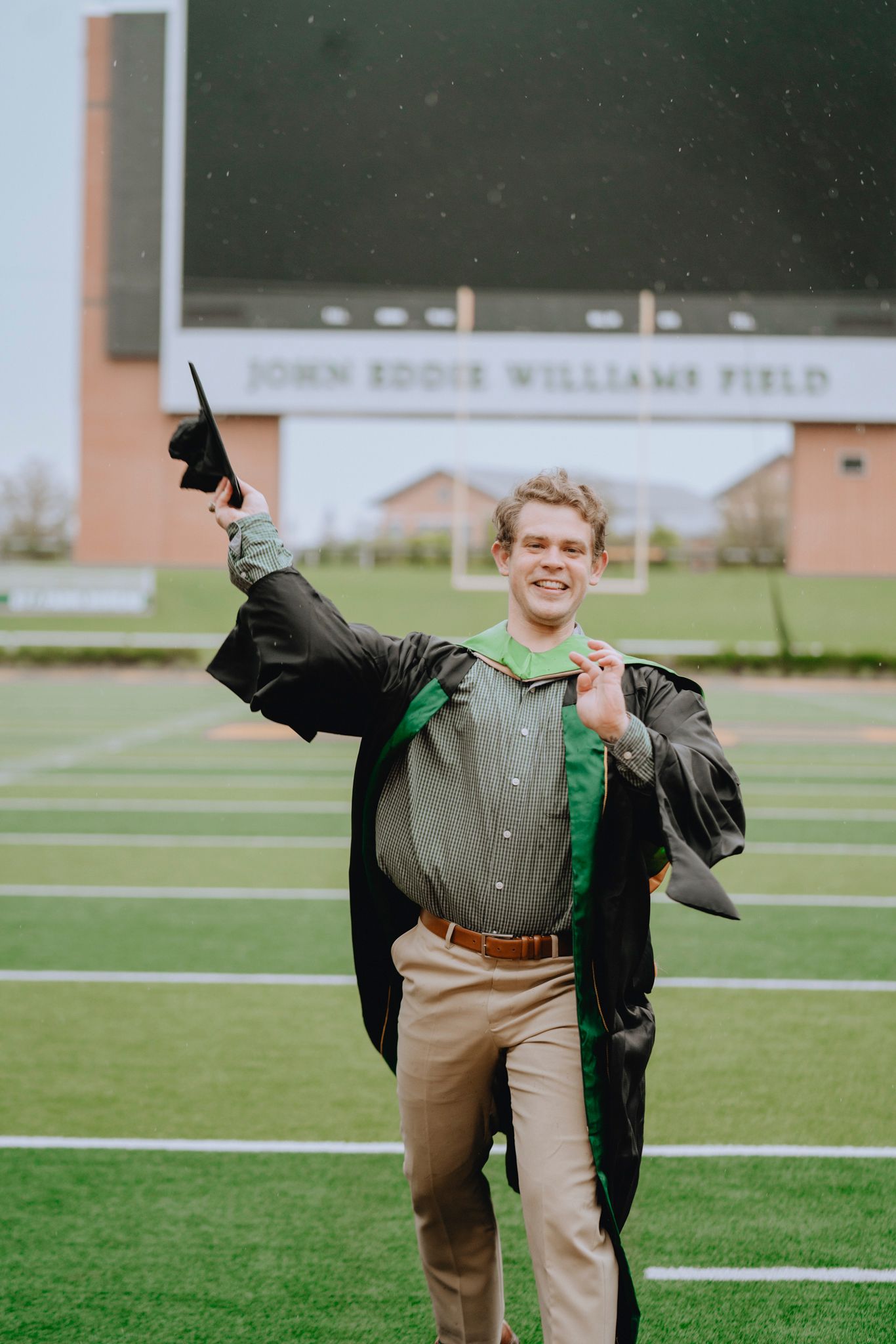 Excited male graduate in black gown celebrates on John Eddie Williams Field football field.