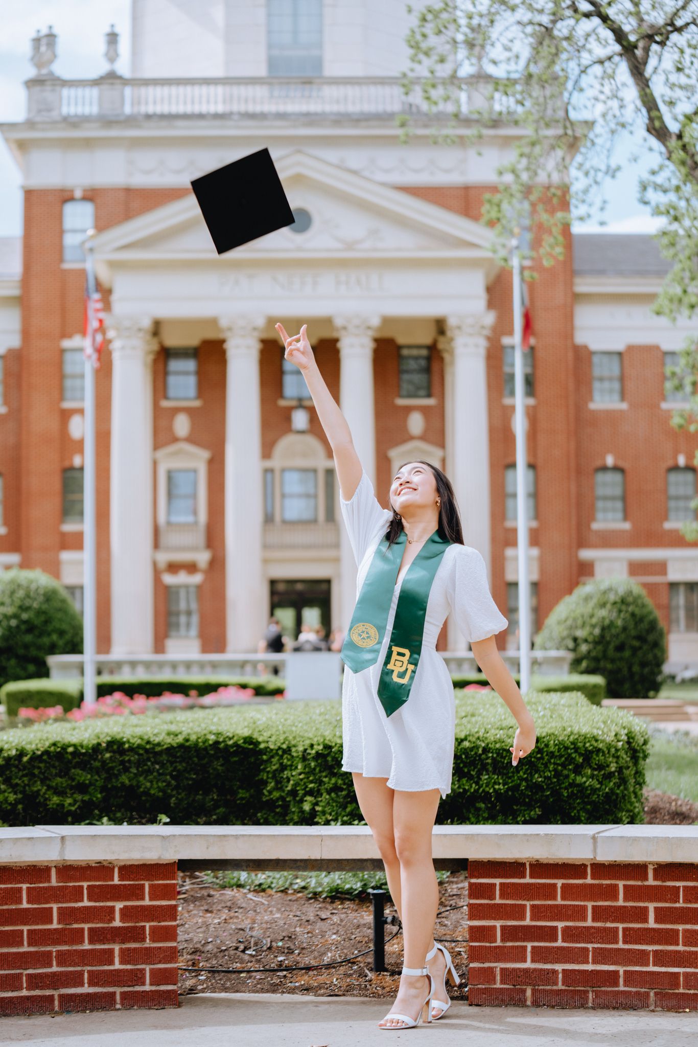 Graduate in white dress tossing cap in front of a classic brick university building.