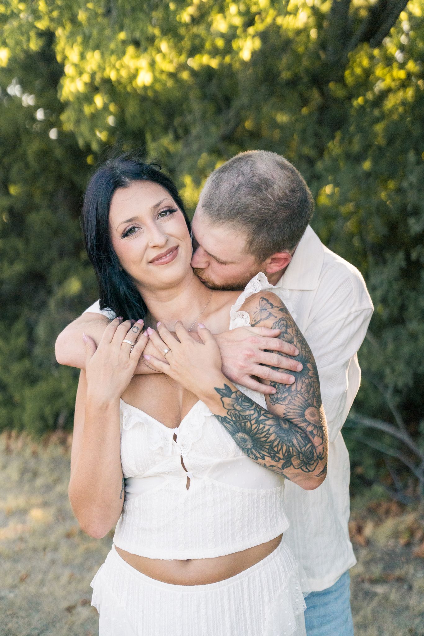 Couple embracing outdoors, man with tattoos kissing woman's cheek while hugging her from behind near green trees.