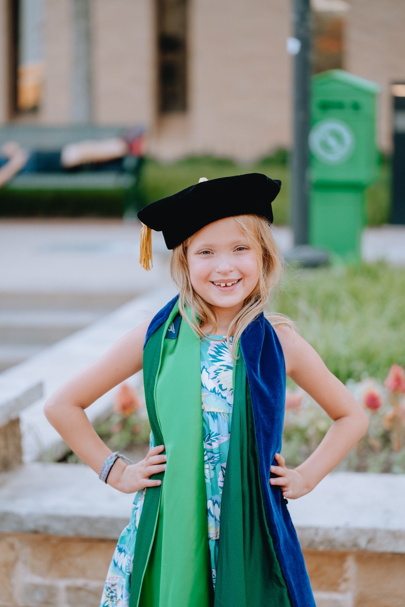 Cheerful young girl posing in green graduation gown and black doctoral cap with hands on hips outdoors.